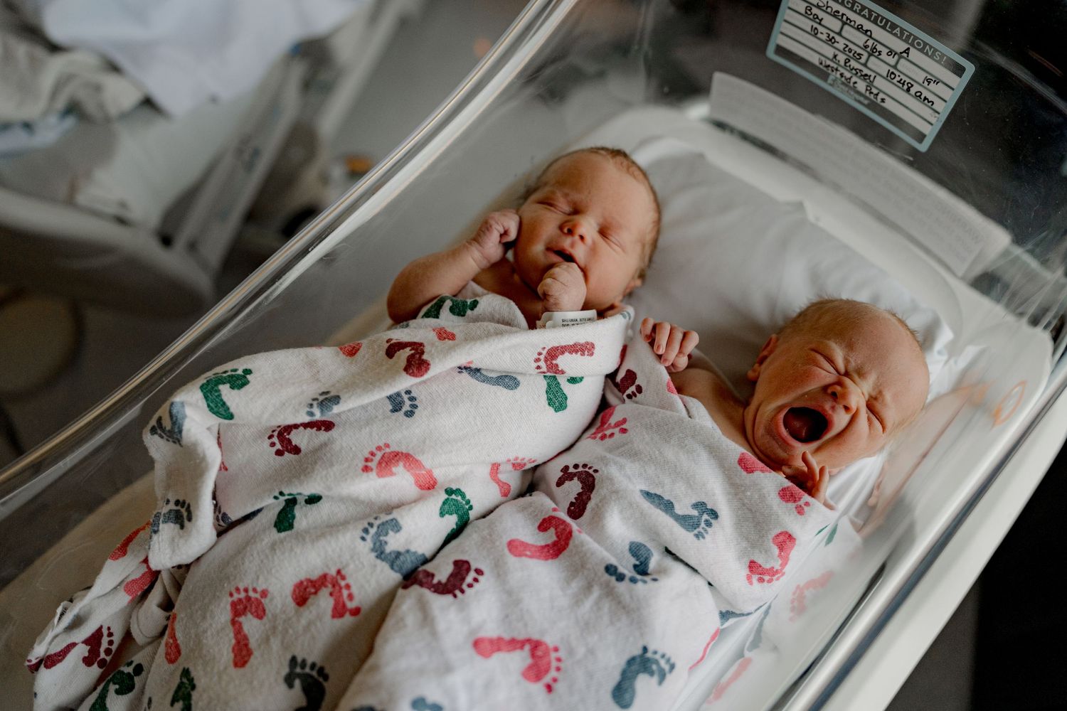 A newborn baby sleeps peacefully in a hospital bassinet wrapped in a white blanket with colorful footprint patterns.