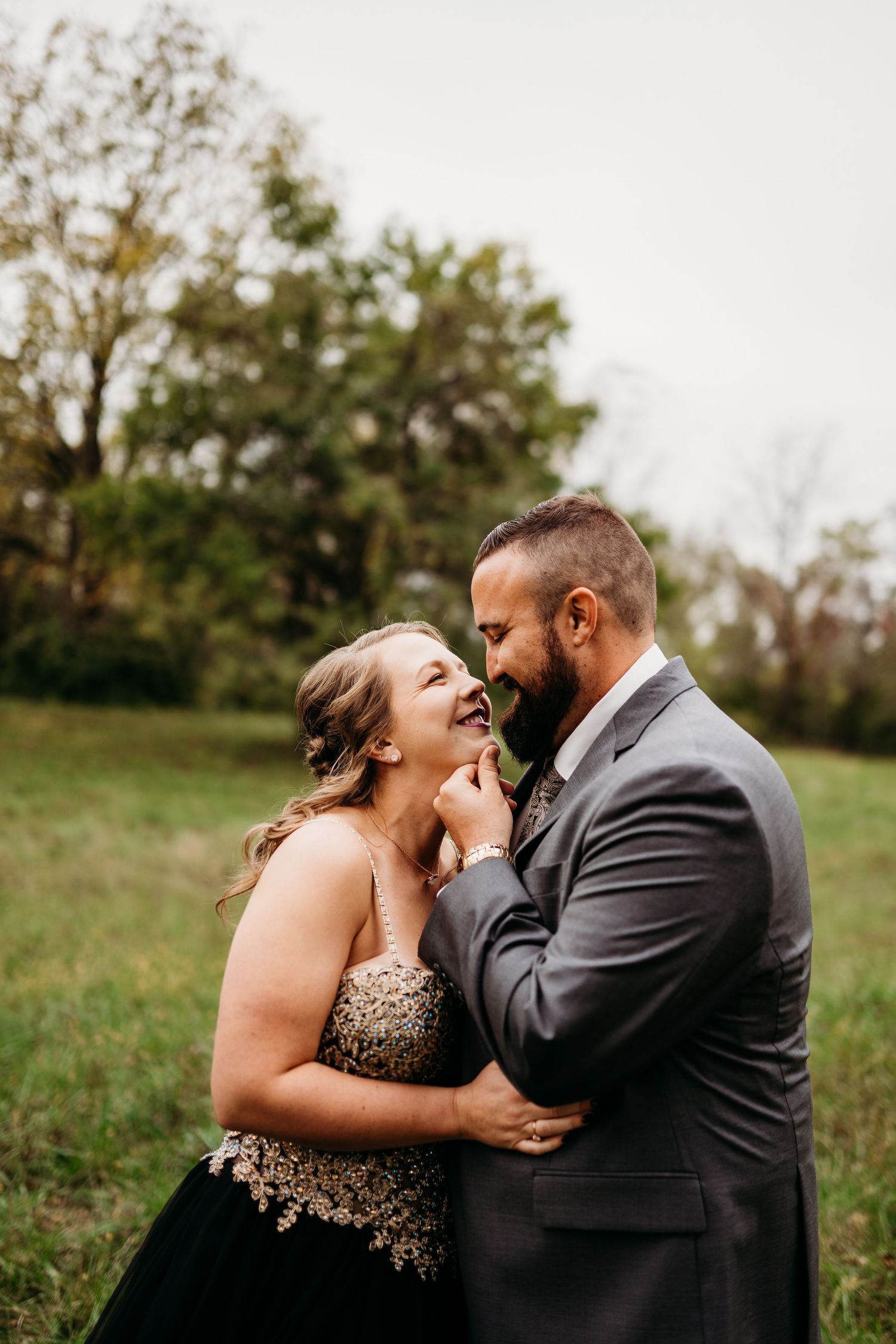 A couple in formal attire embraces and laughs together in a grassy outdoor setting with trees in the background.