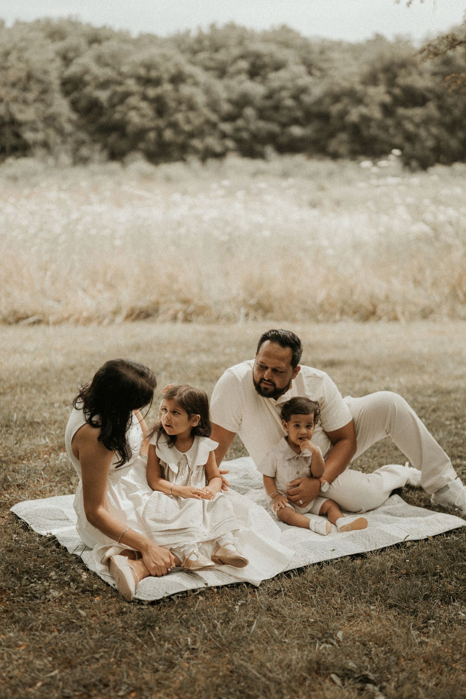 Family of four in white outfits enjoying a picnic on a blanket in a scenic meadow with lush greenery.