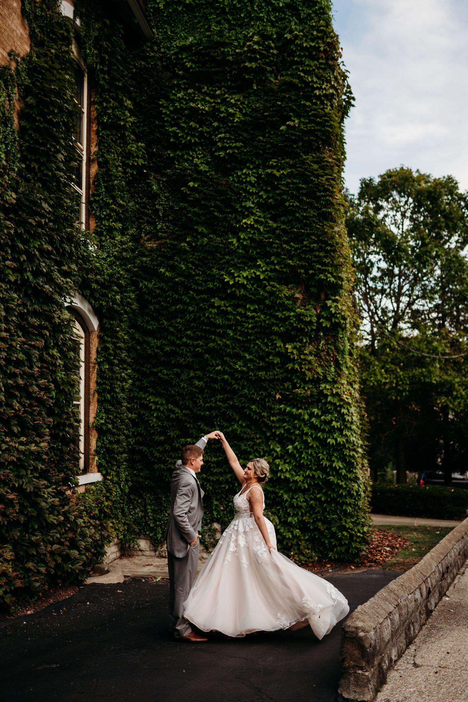 A romantic wedding couple dances and twirls together against an ivy-covered wall in an elegant outdoor setting.