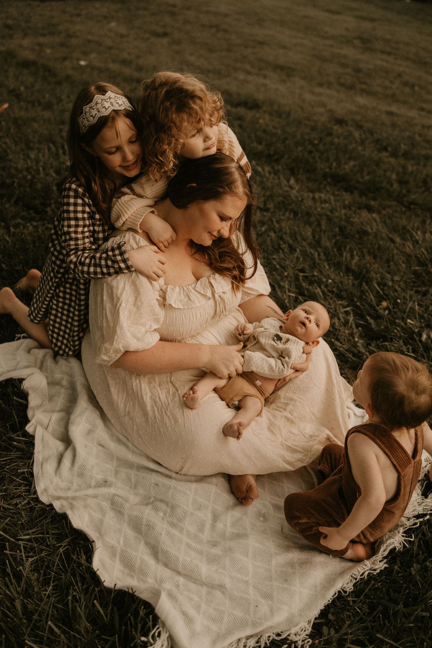 A mother sits on a white blanket in a field cradling her newborn, surrounded by her three young children in warm tones.