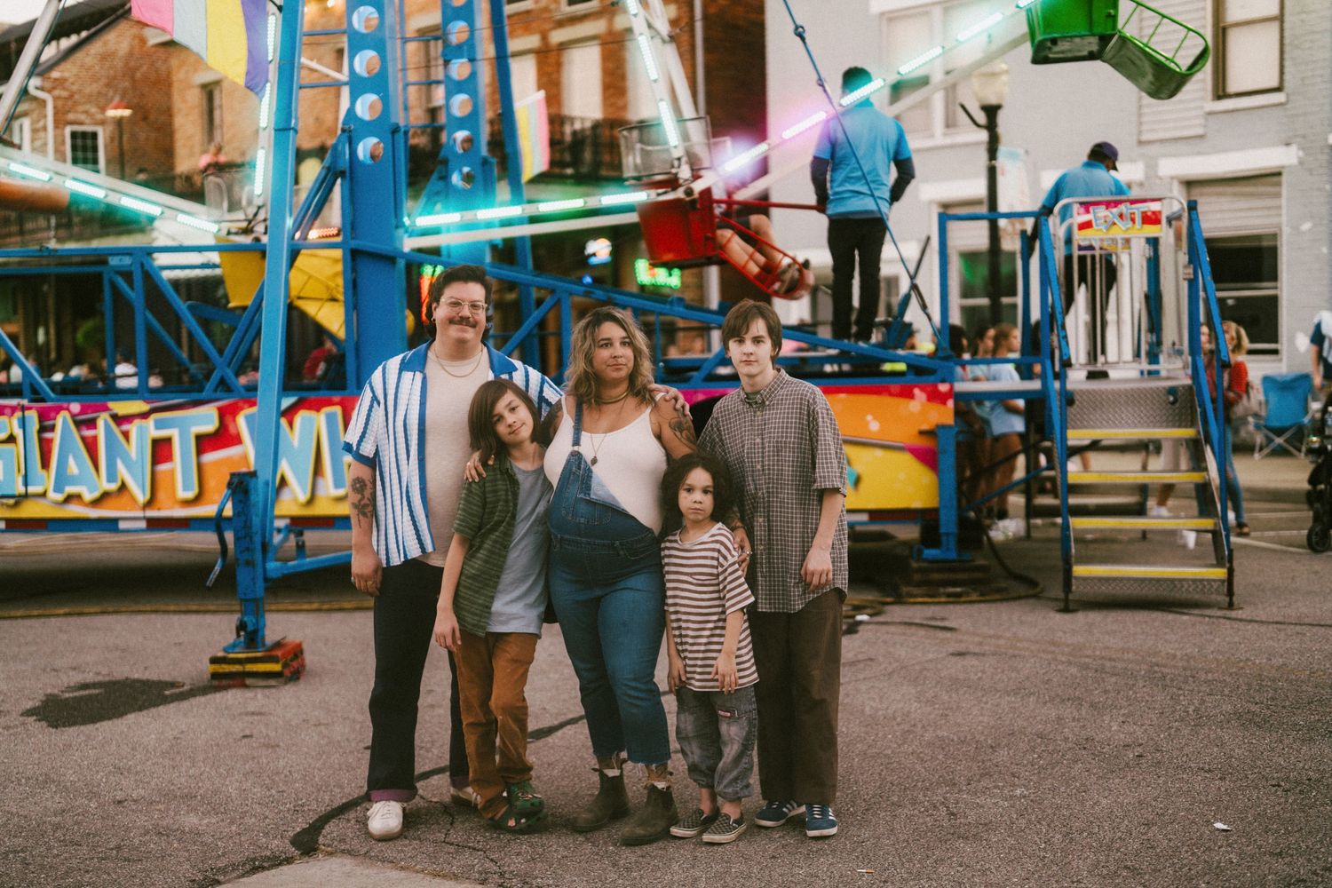 Family posing together at a colorful carnival fairground with rides and lights in the background.