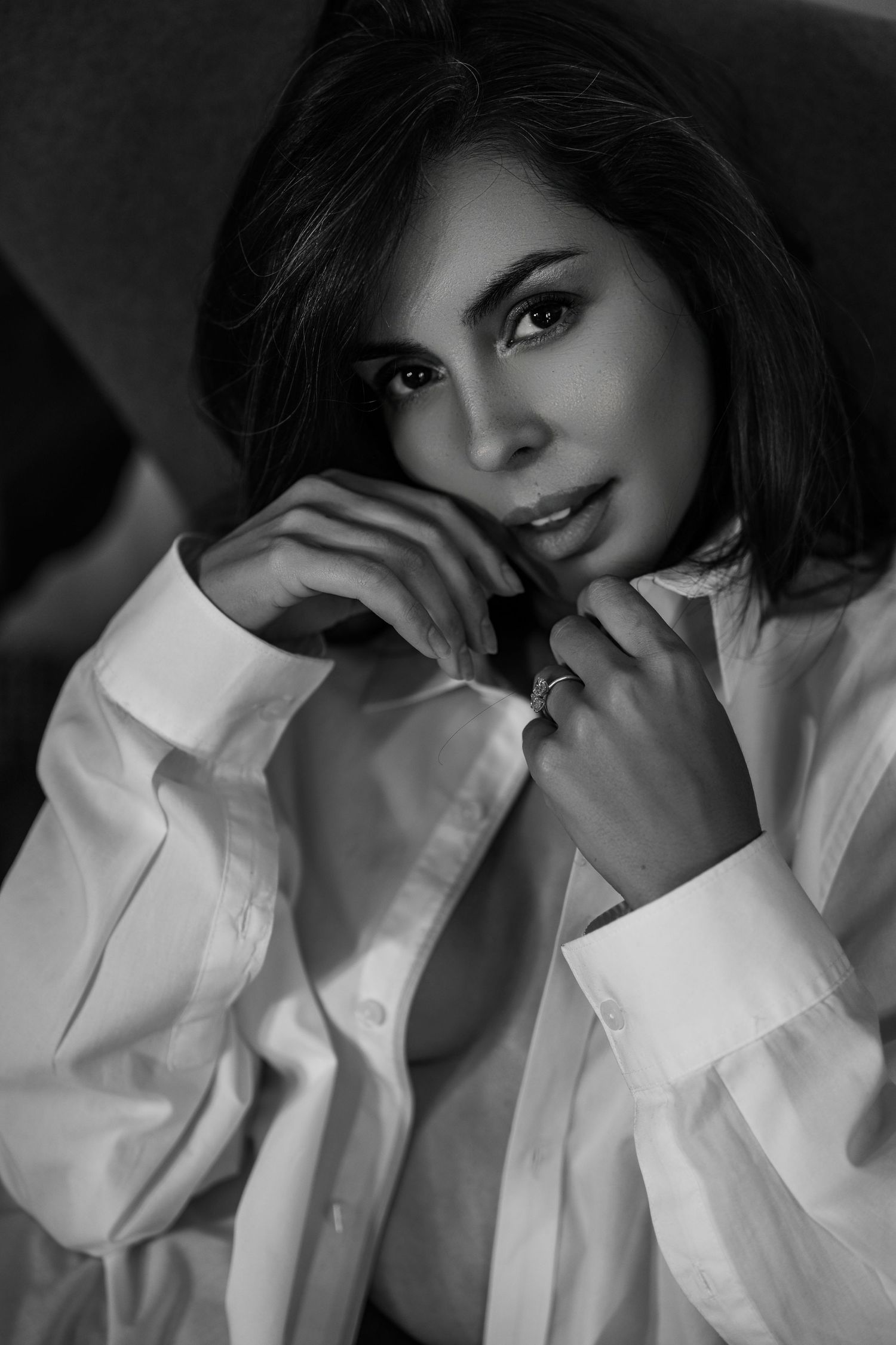 Black and white portrait of a person wearing an oversized white button-up shirt while posing with hands near face.