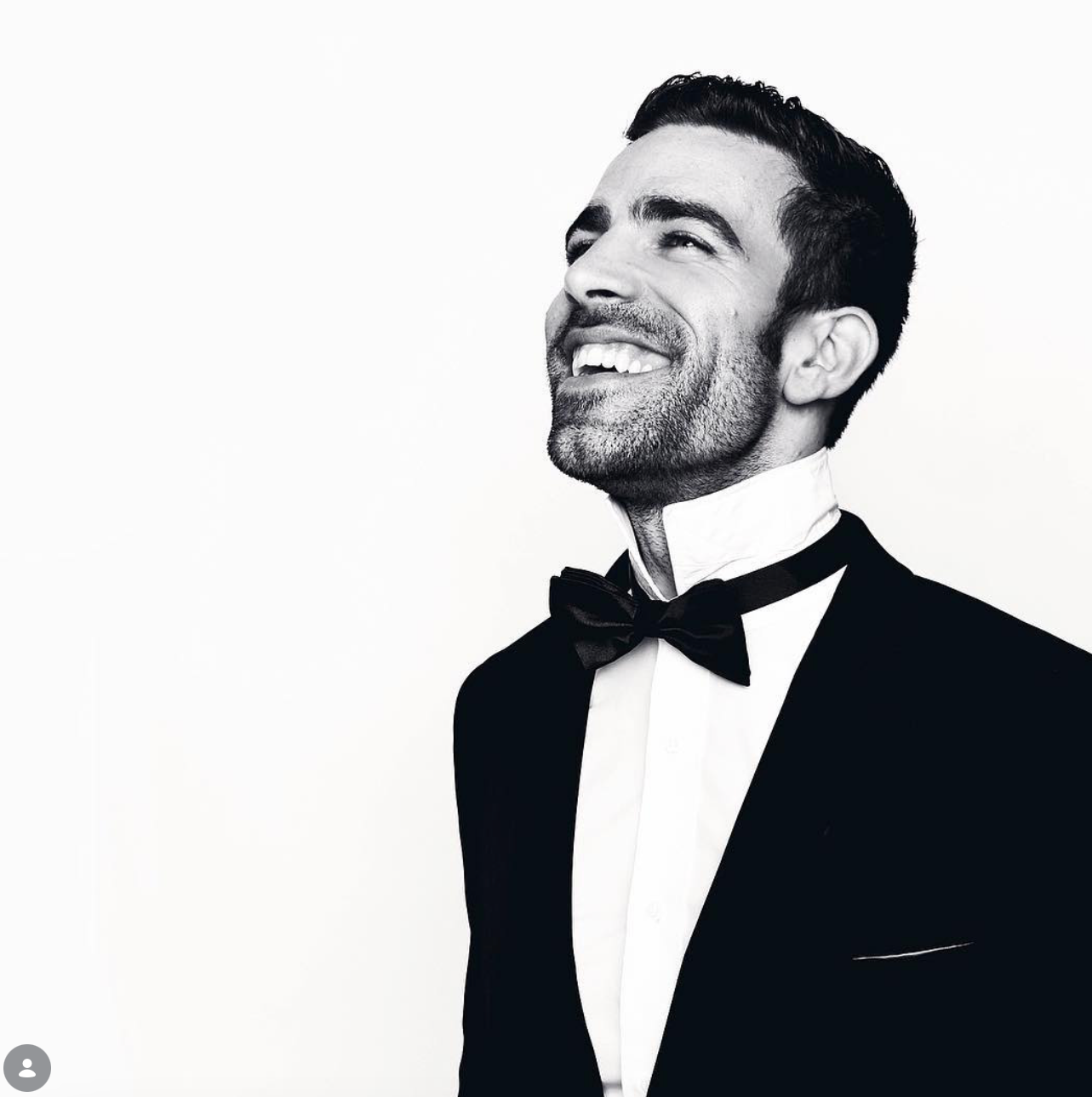 Black and white portrait of a smiling bearded man in a tuxedo and bow tie, gazing upward against a plain white background.