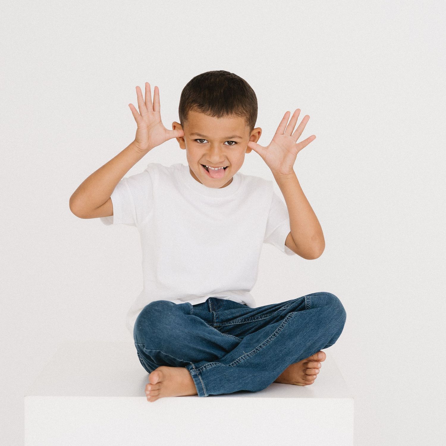 Young person in white t-shirt and jeans sitting cross-legged on white background making playful expressions.