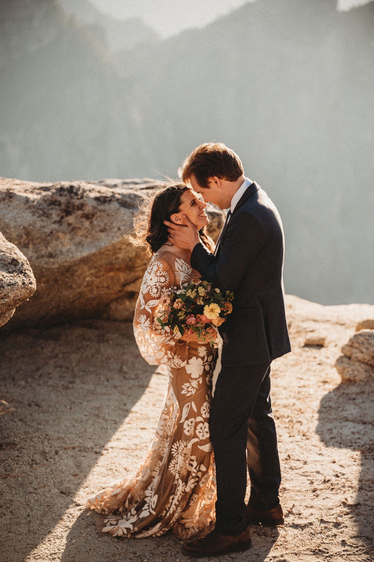 A couple in wedding attire shares an intimate moment on a rocky cliff during golden hour sunset.