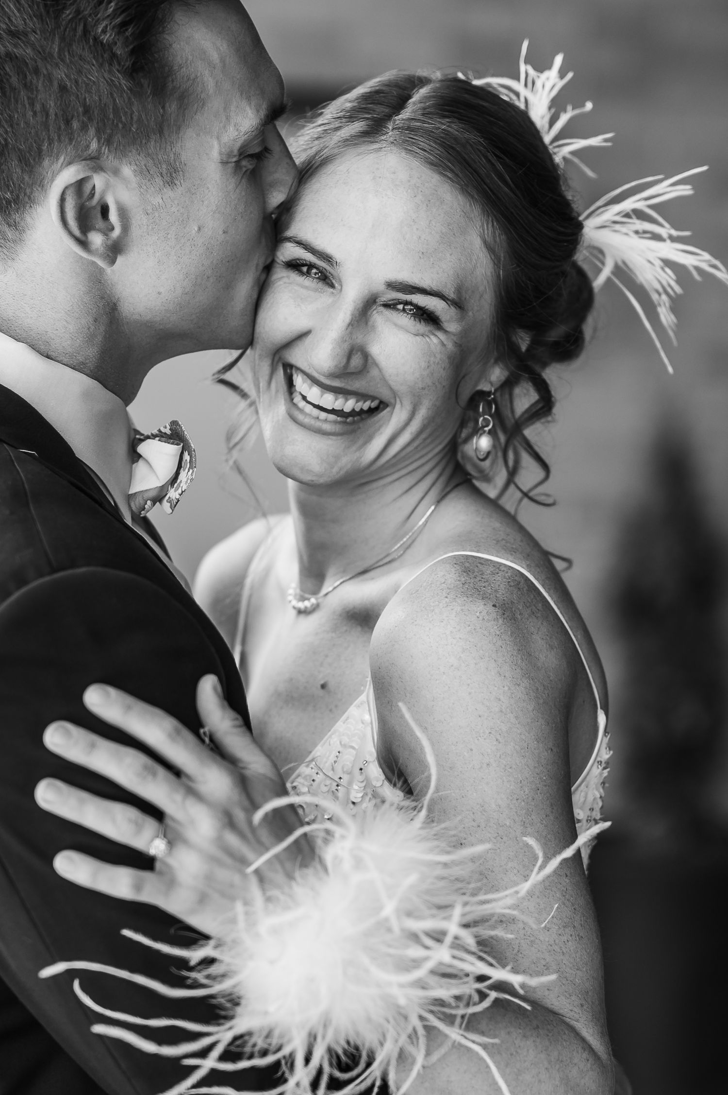 Black and white wedding photo of a smiling bride in feathered dress being embraced by groom in suit.