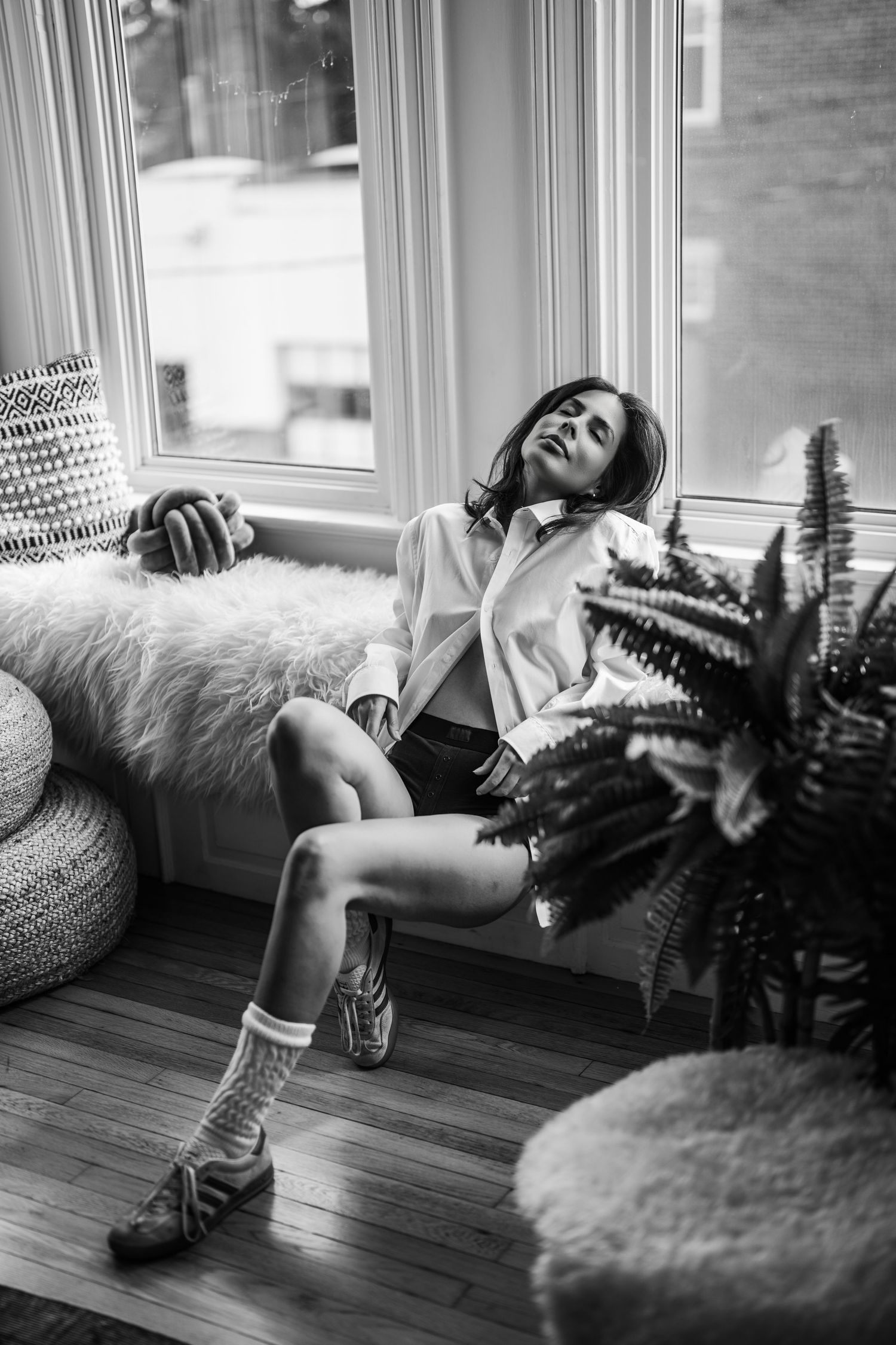 A black and white photo series showing a person lounging by a window with ferns and a fluffy rug on a wooden floor.