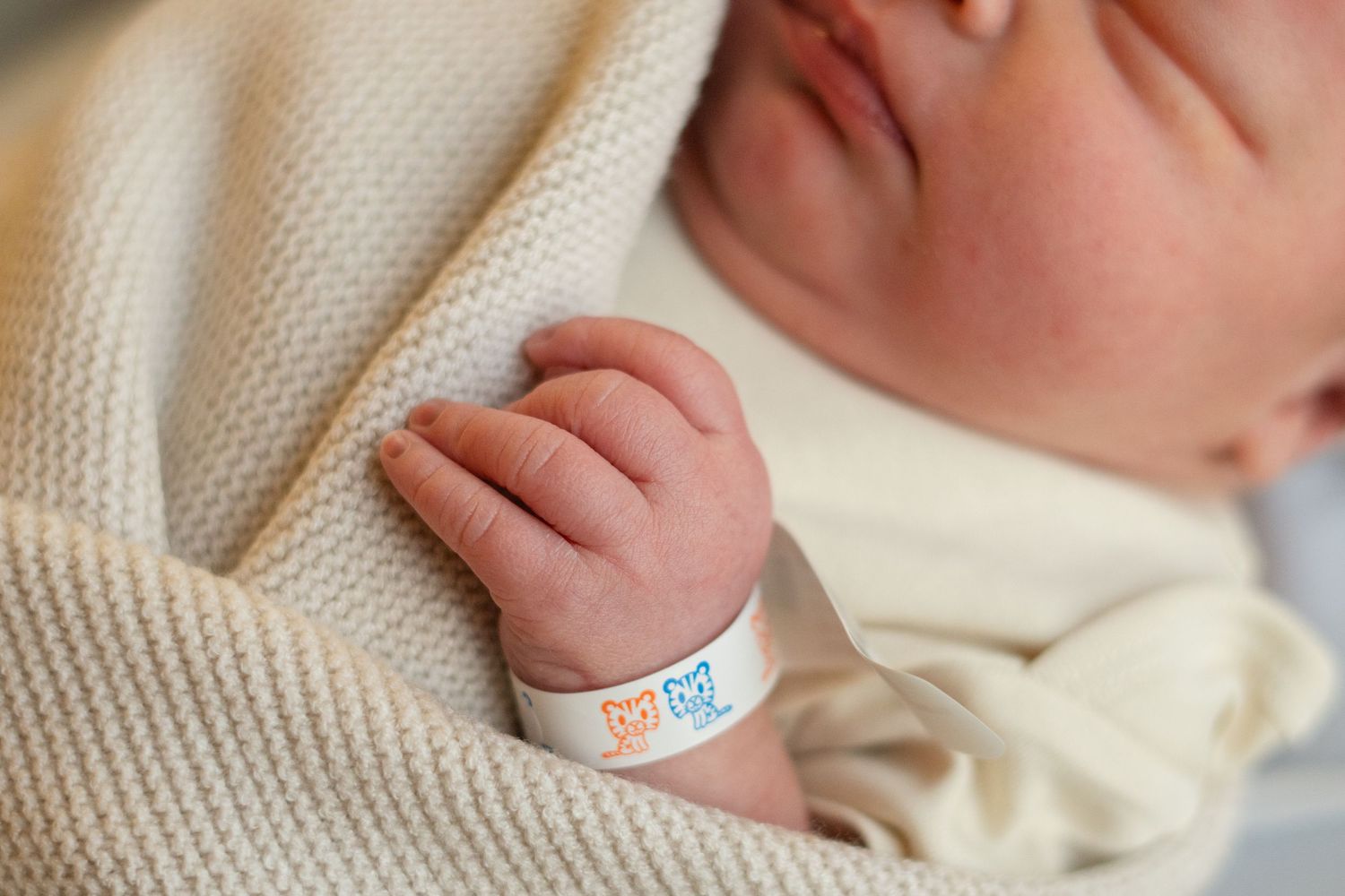 A tiny newborn baby hand with hospital identification bracelet rests on soft white blanket.