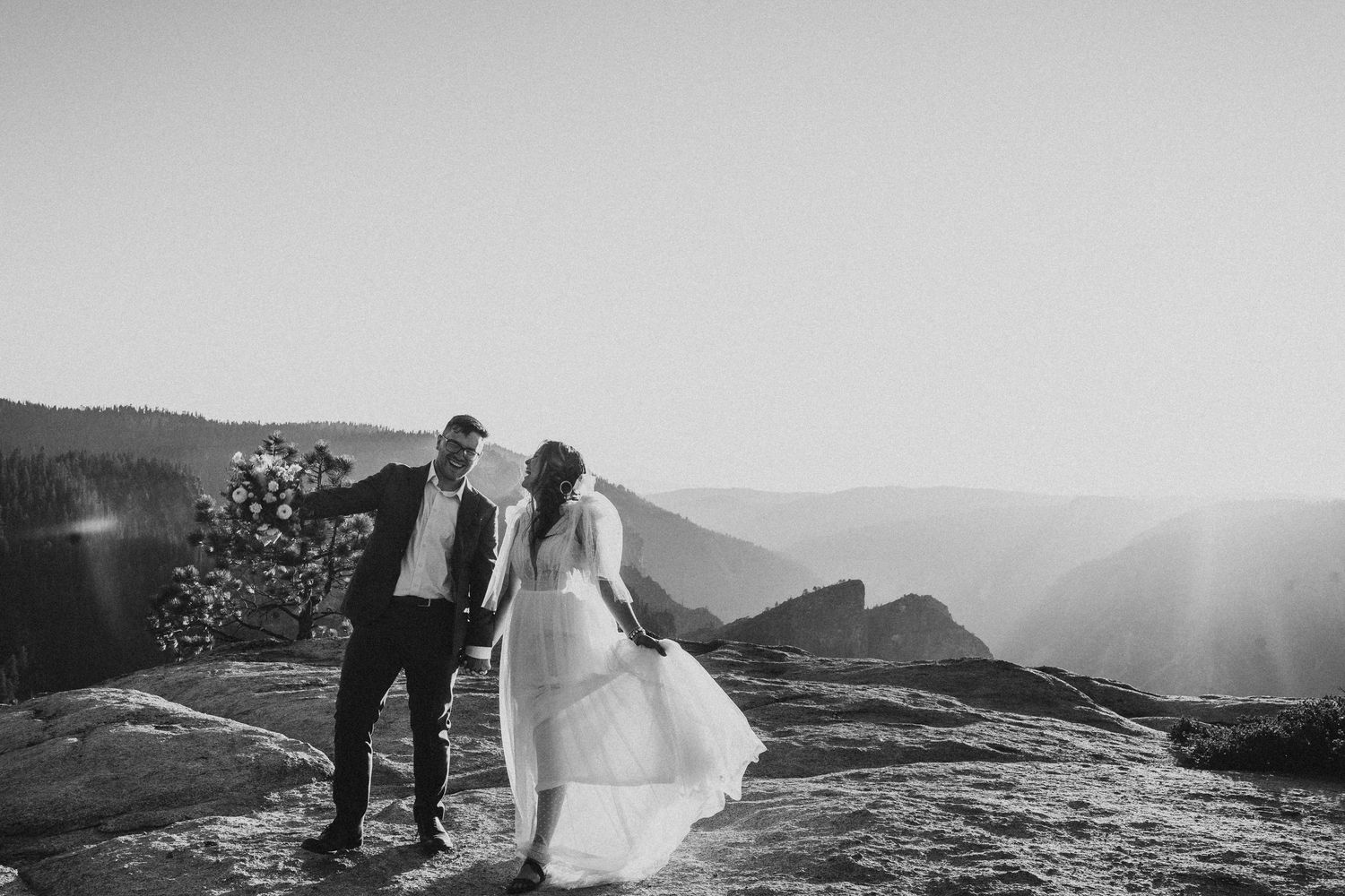 A couple in wedding attire poses together on a scenic mountain overlook at sunset creating a romantic silhouette.