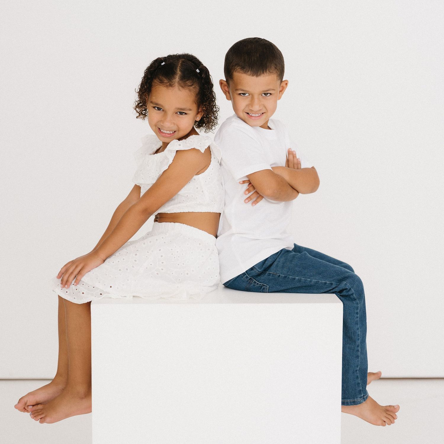 Two children in white tops and jeans sit and play together on a white surface in a minimalist setting.