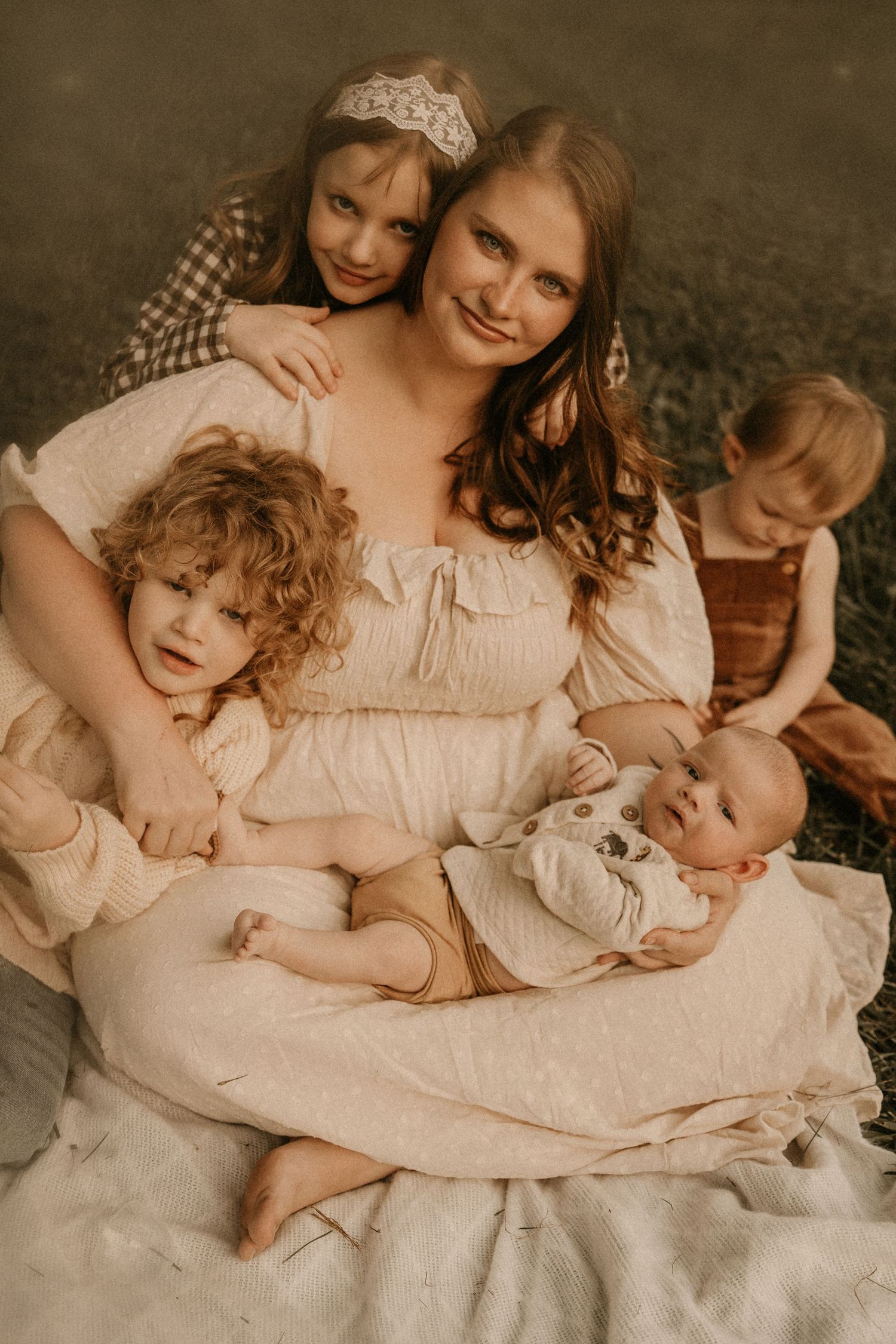 A mother in a white dress sits outdoors on a blanket surrounded by her four young children in a warm family portrait.