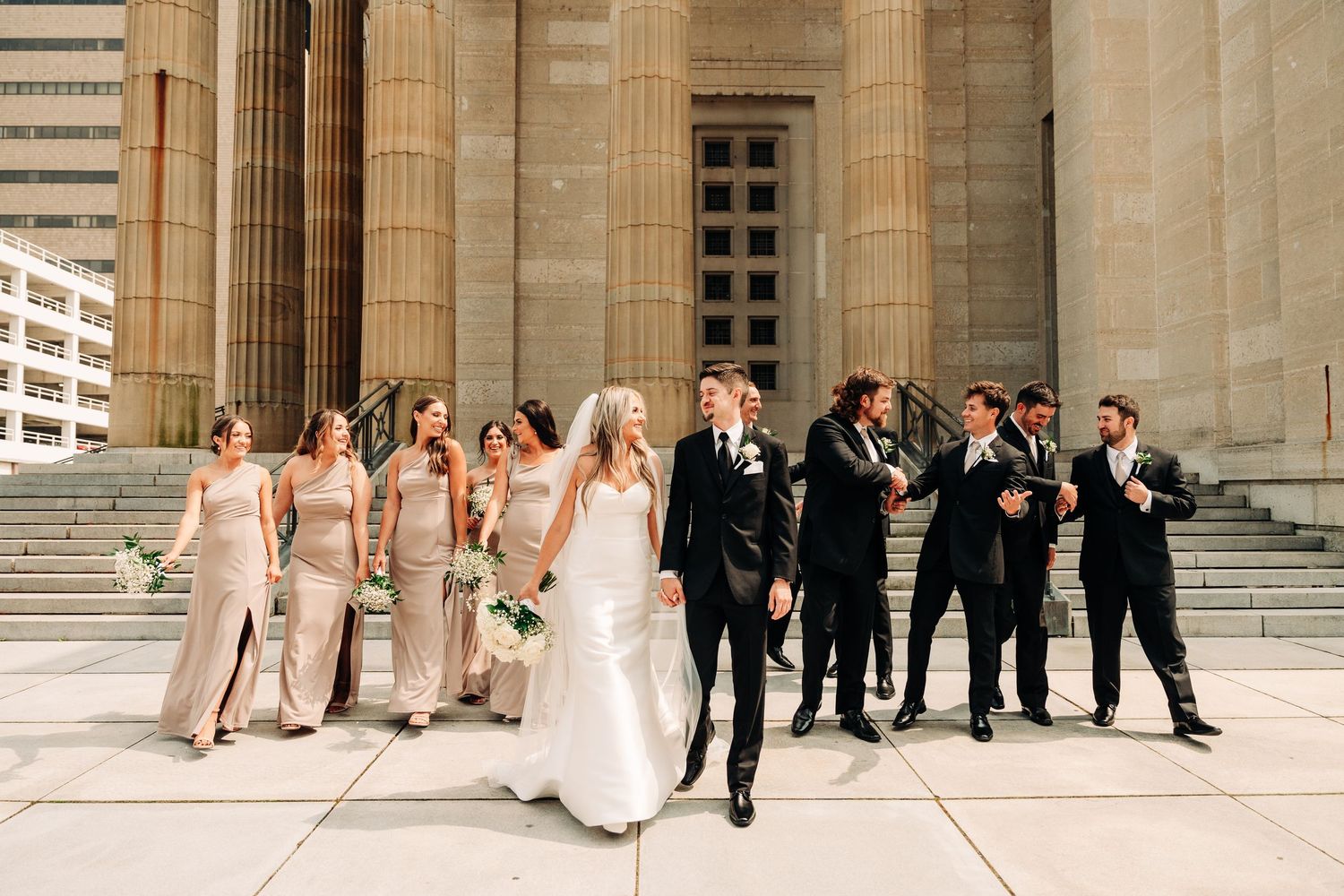 Wedding party poses in sequence of candid shots along elegant stone building columns during outdoor celebration.
