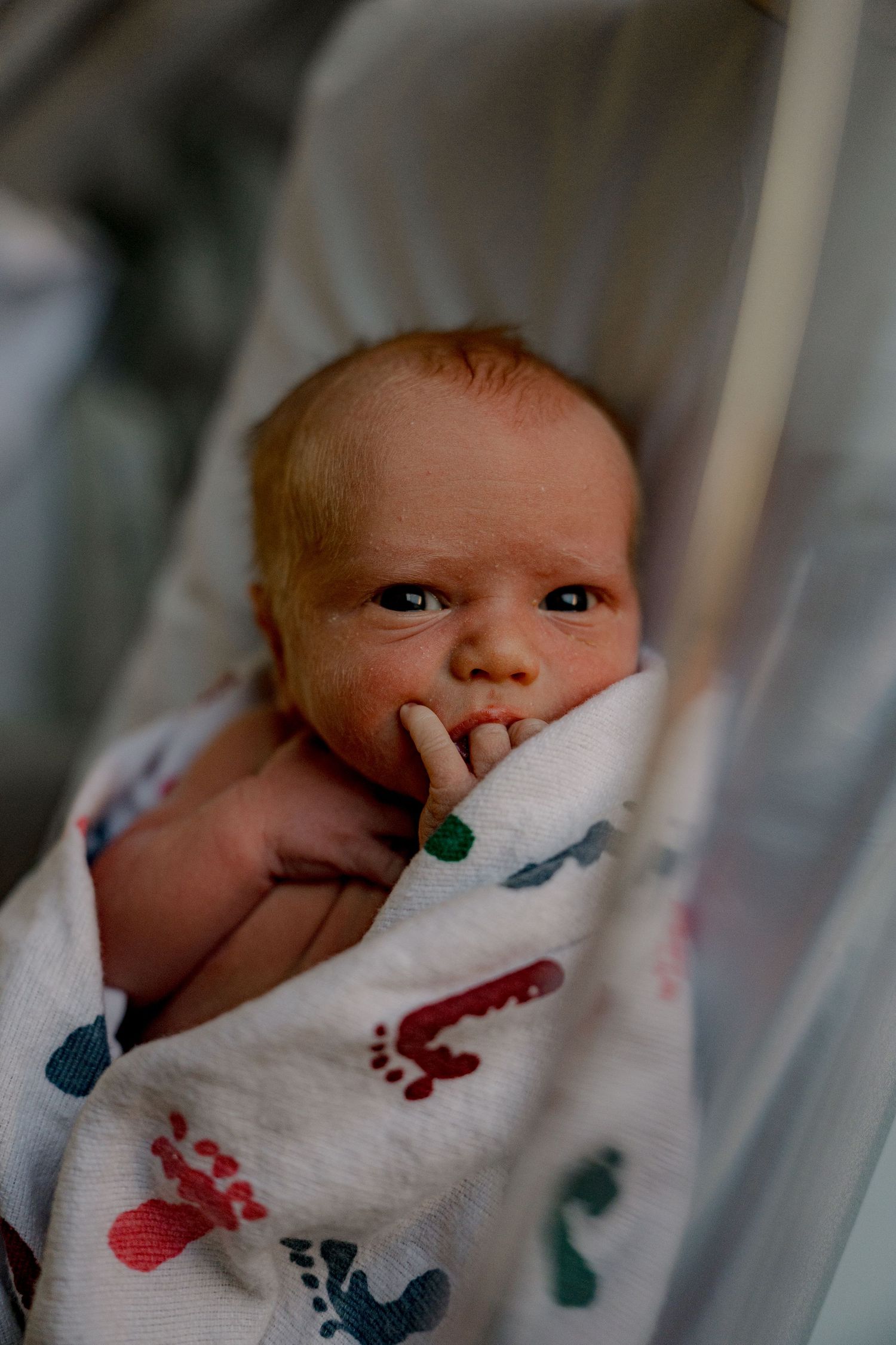 A newborn baby wrapped in a white blanket with colorful handprint patterns gazes upward while resting.