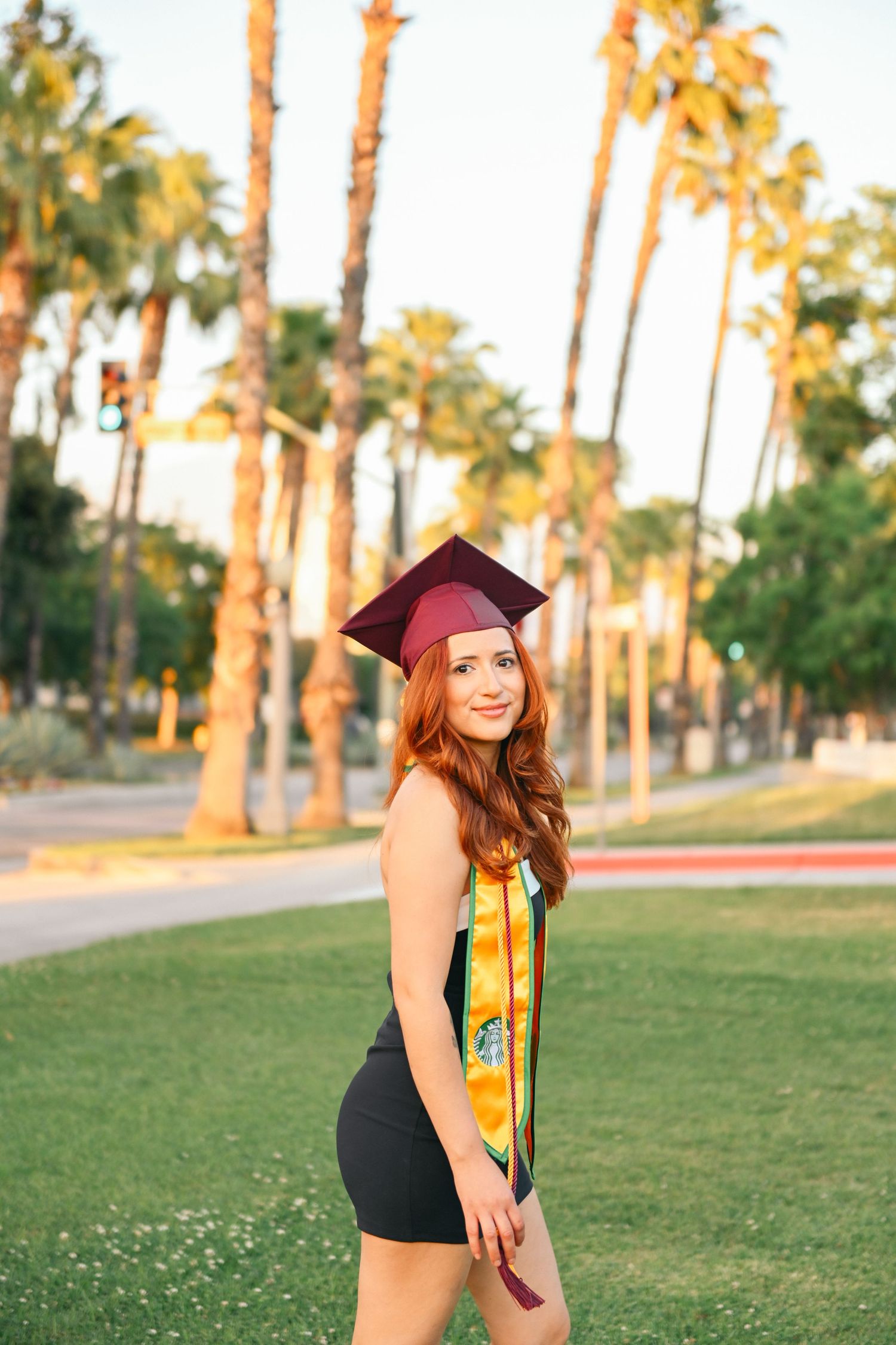 Graduate in black dress and maroon cap poses on lawn with palm trees during golden hour.