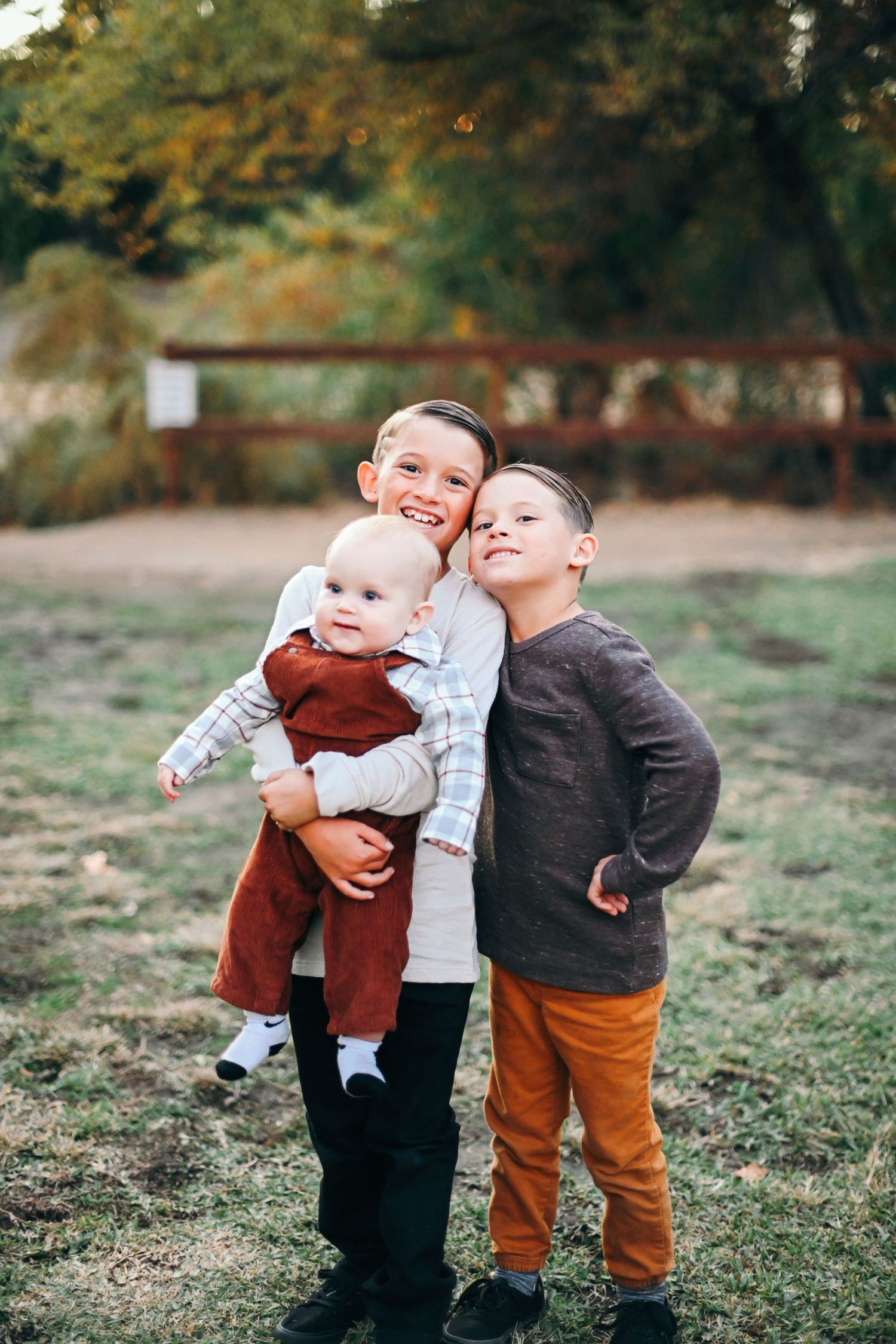 Three young children pose together outdoors during autumn, with fallen leaves in background.