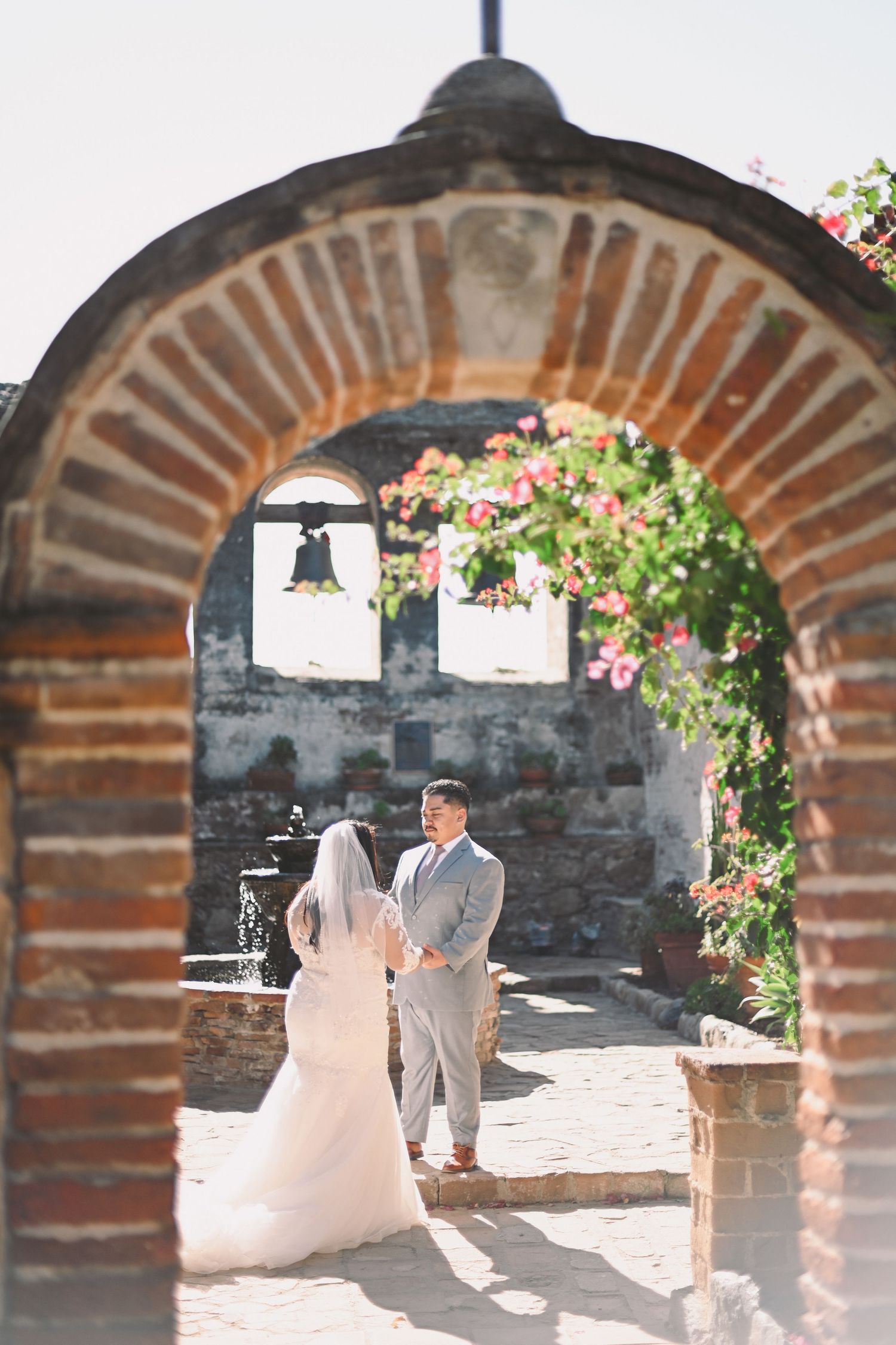 Wedding couple standing under a beautiful brick archway adorned with pink flowers.