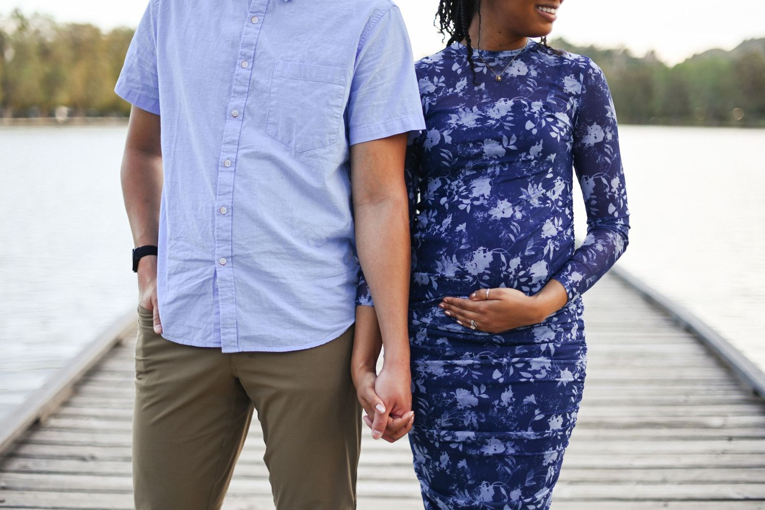 A couple stands on a wooden dock, wearing blue clothing for their maternity photo.