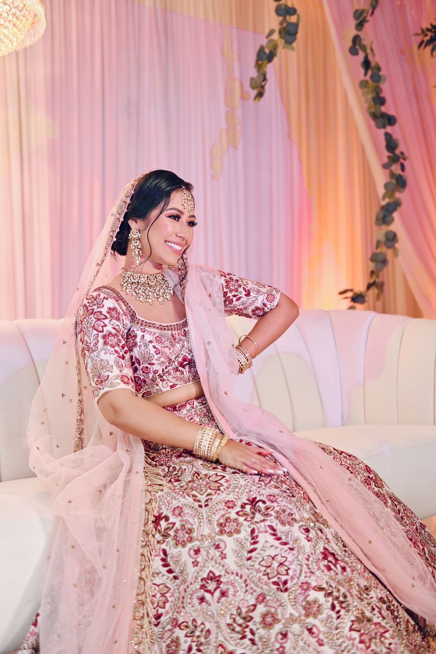 Traditional South Asian bride in pink embellished lehenga sitting on white couch.