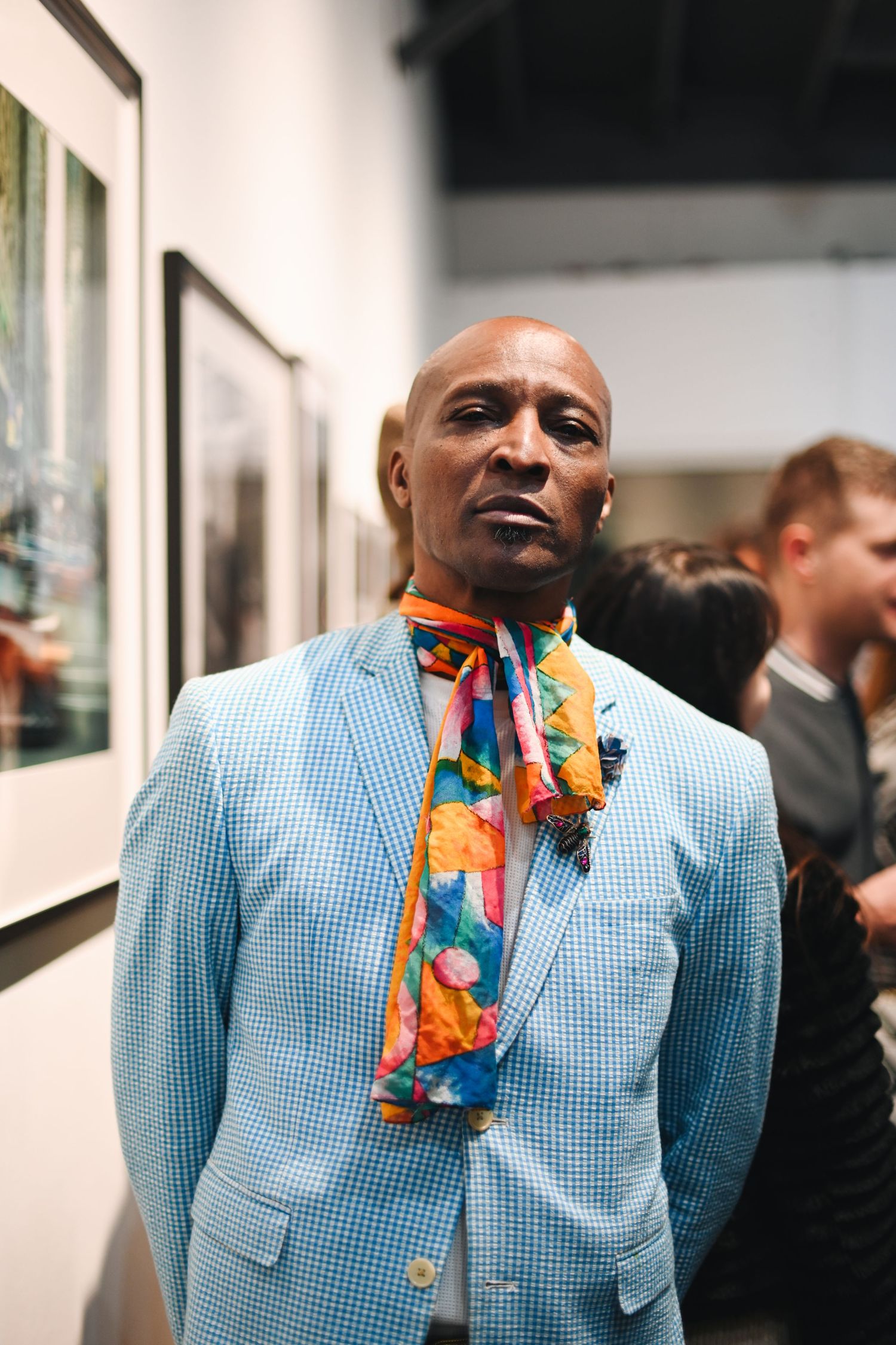Stylish gentleman in light blue checkered suit and colorful scarf standing against gallery wall.
