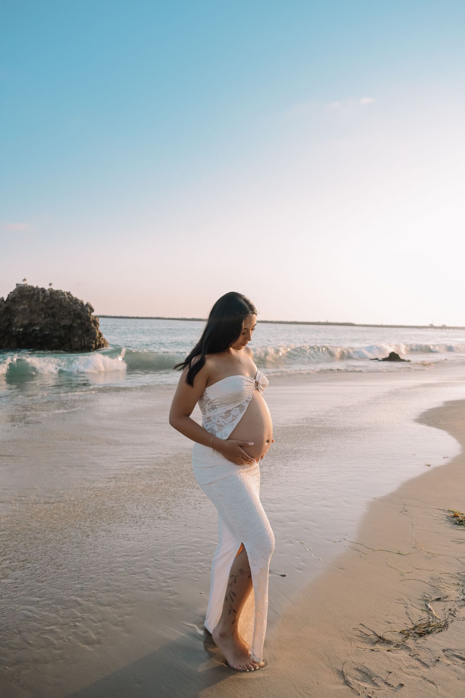 Woman in white dress poses for maternity photo on beach at sunset with waves and rocks in background.