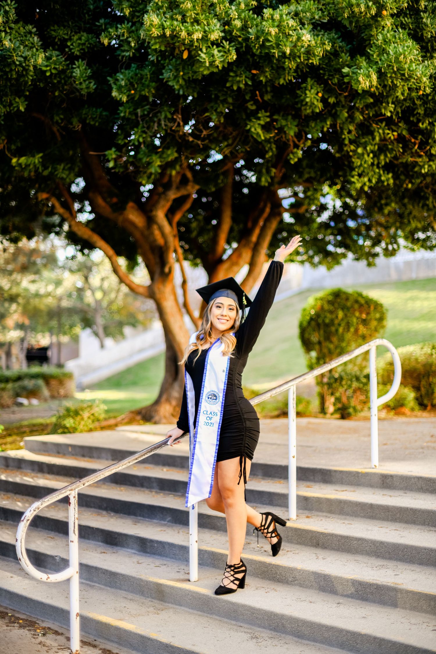 Graduate in black dress and sash poses joyfully on outdoor steps with large tree in background.