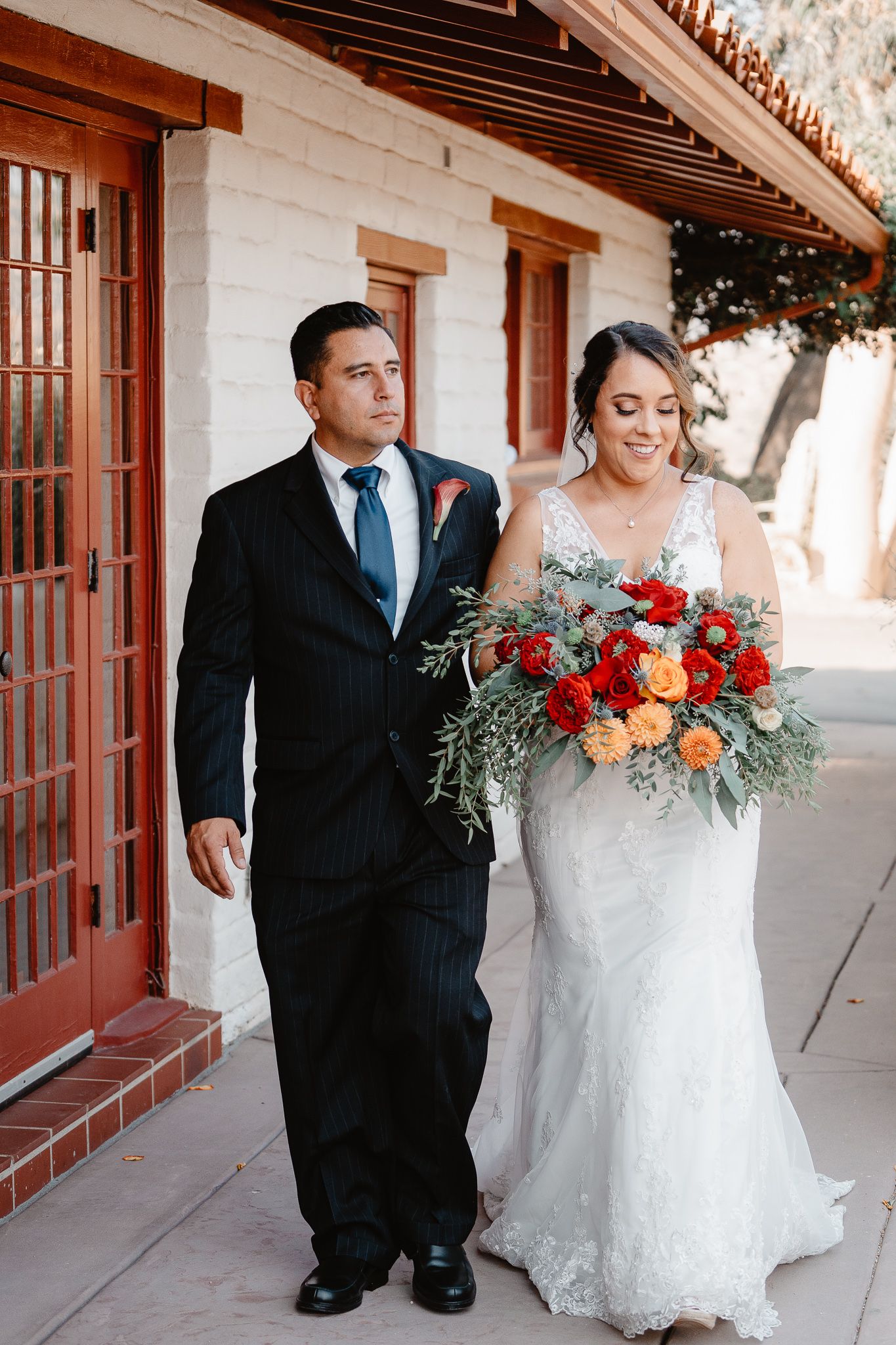 Wedding couple walking along Spanish colonial building with vibrant red and orange bouquet.