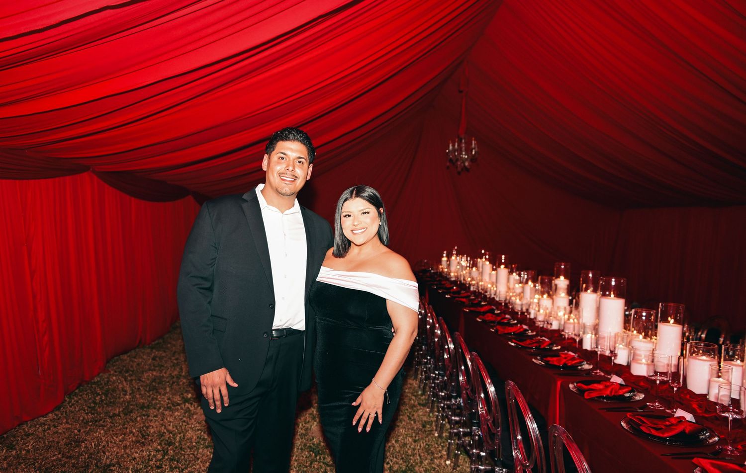 Formal couple standing in front of long table with red decor and candlelight.