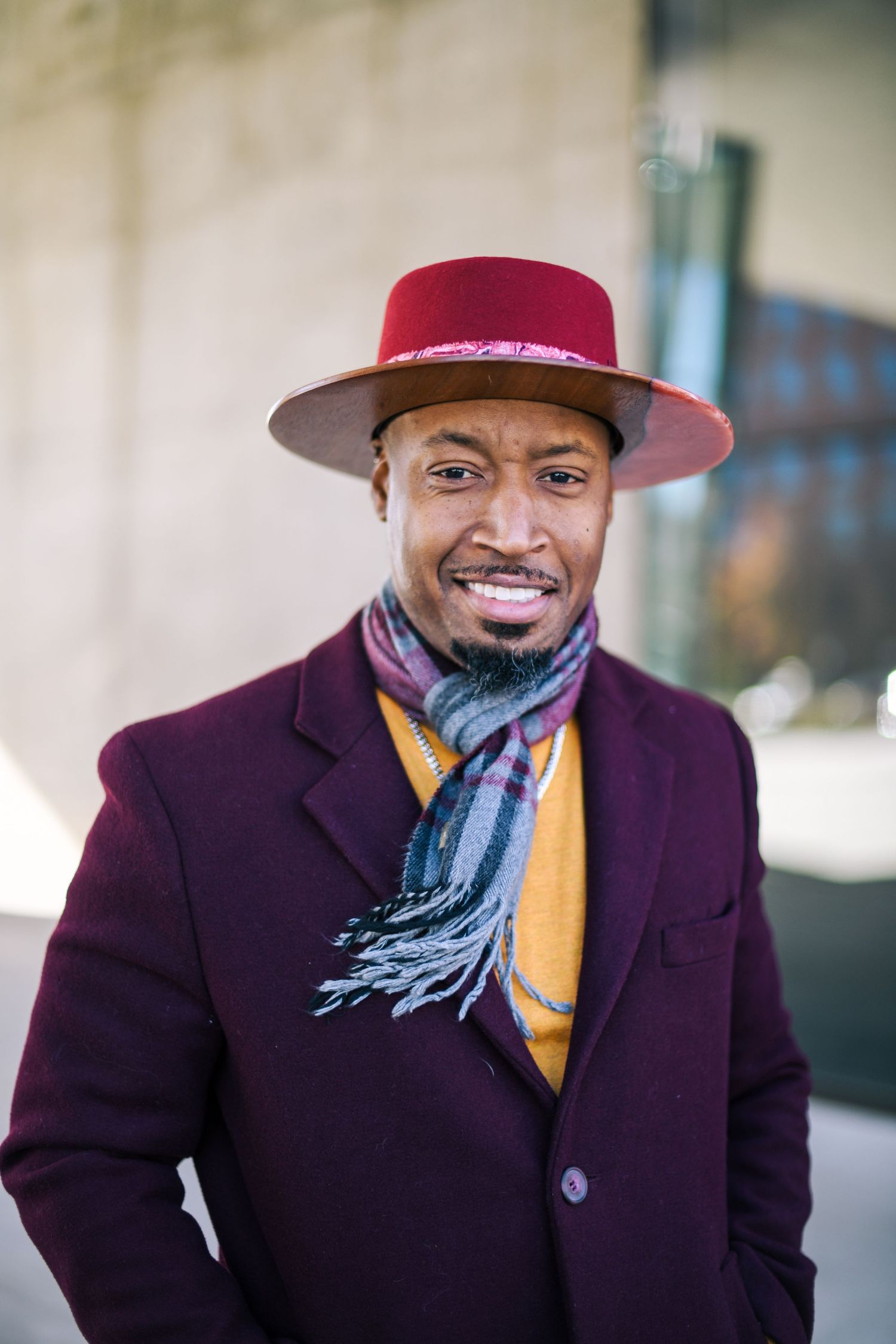 Stylish person in burgundy coat and red hat with scarf against urban background.