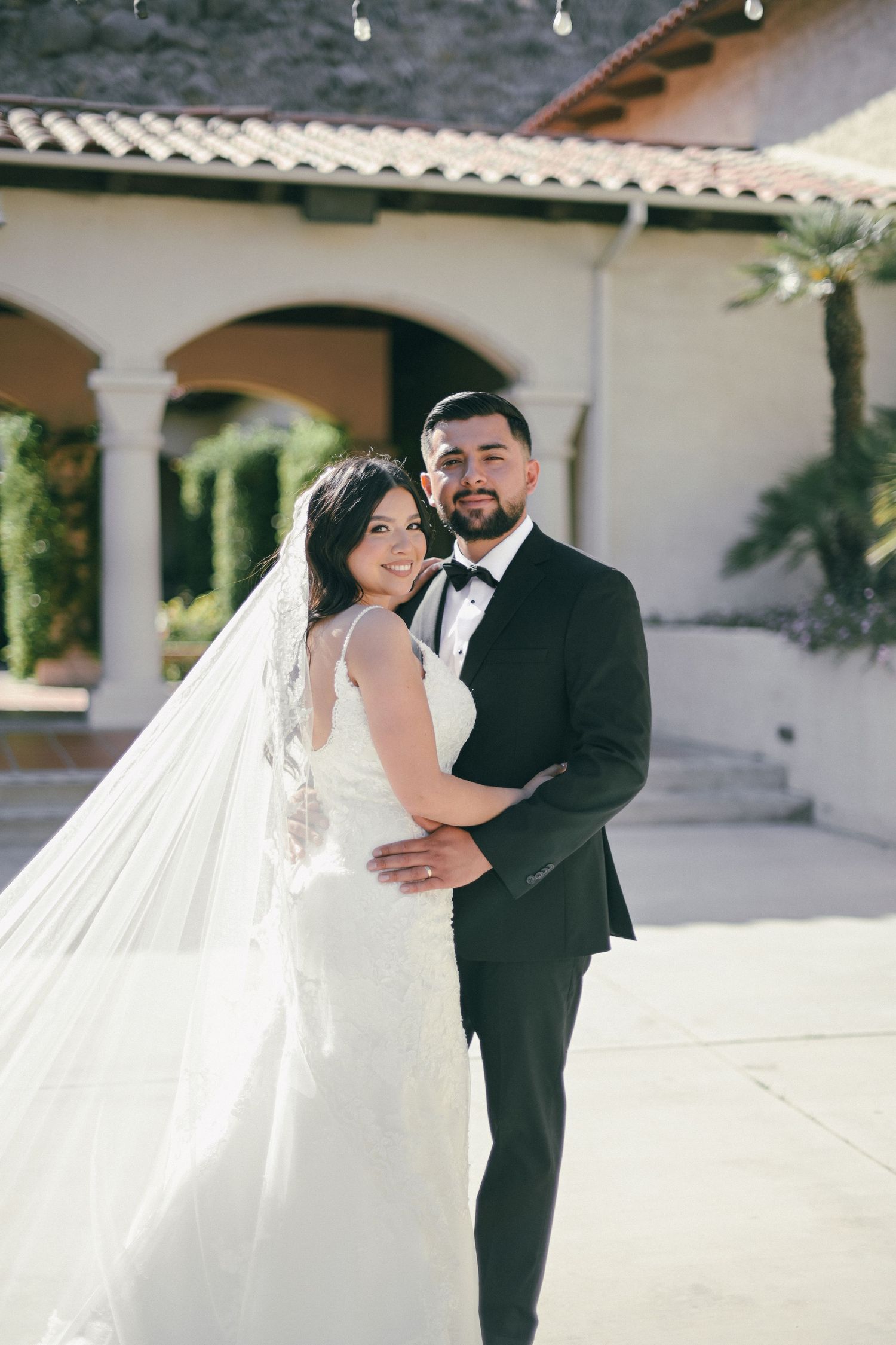 Wedding couple poses in front of Mediterranean style building with arches and terra cotta roof.