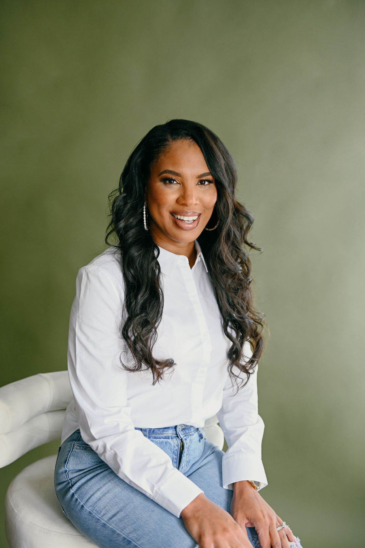 Professional portrait of person in white sweater and jeans sitting on light colored chair.
