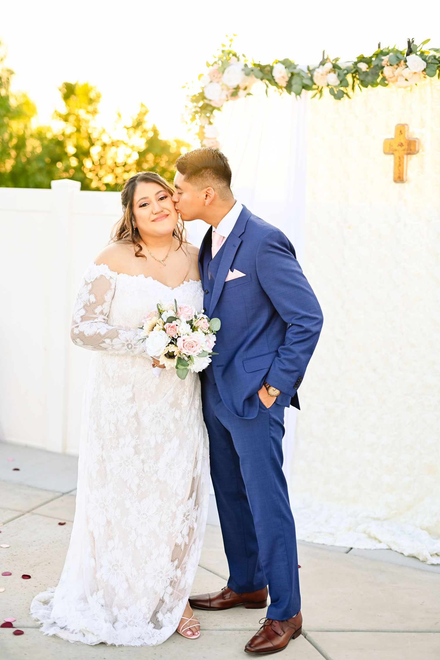 A romantic moment between newlyweds in front of a white floral arch during golden hour.