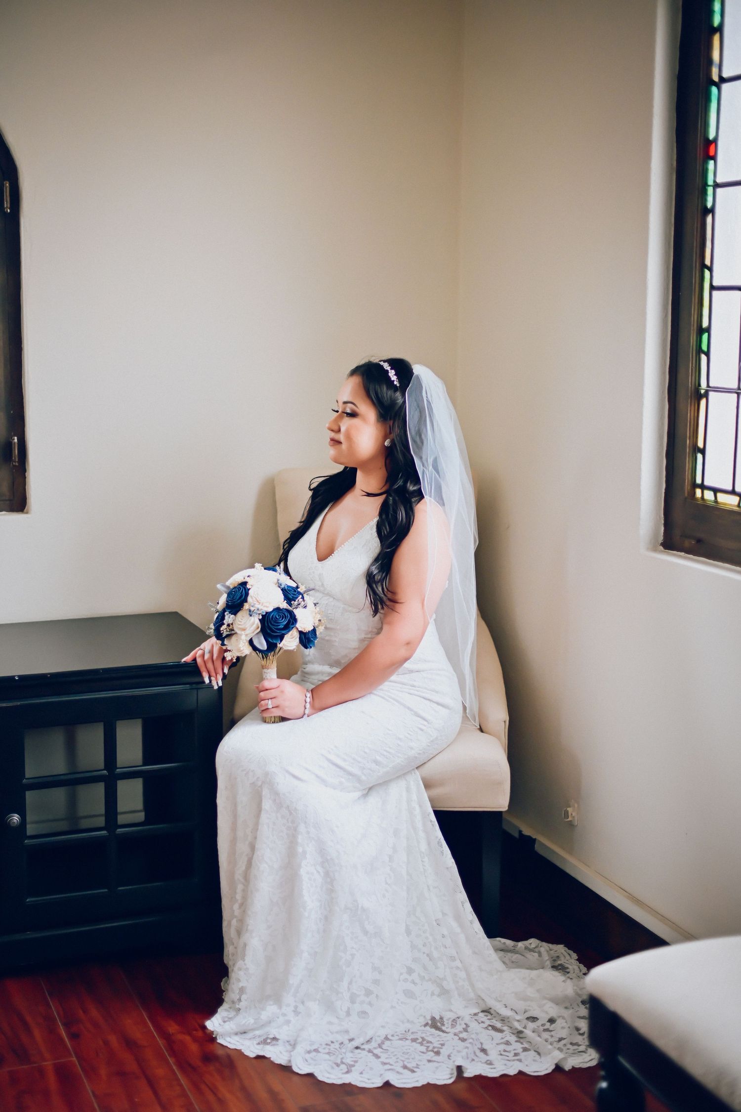 A bride in a lace wedding gown sits with a bouquet by a stained glass window.