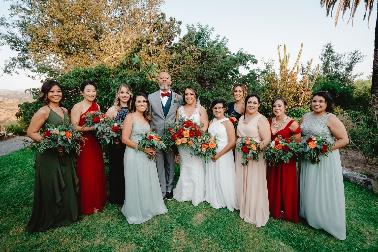 Wedding party poses outdoors in mix of burgundy and neutral dresses with lush fall floral bouquets.