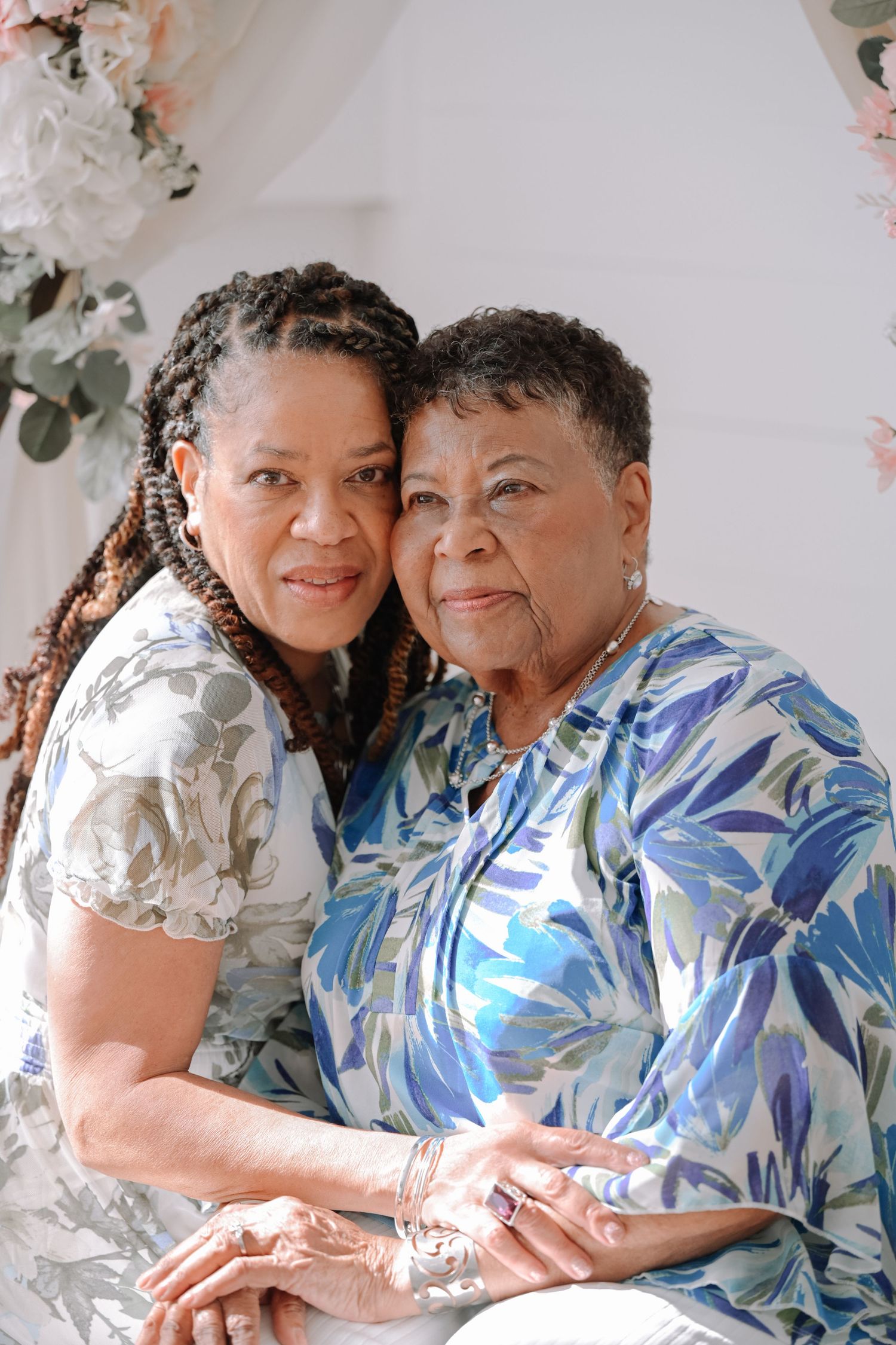 Two women wearing floral and white outfits embrace warmly during indoor portrait session.