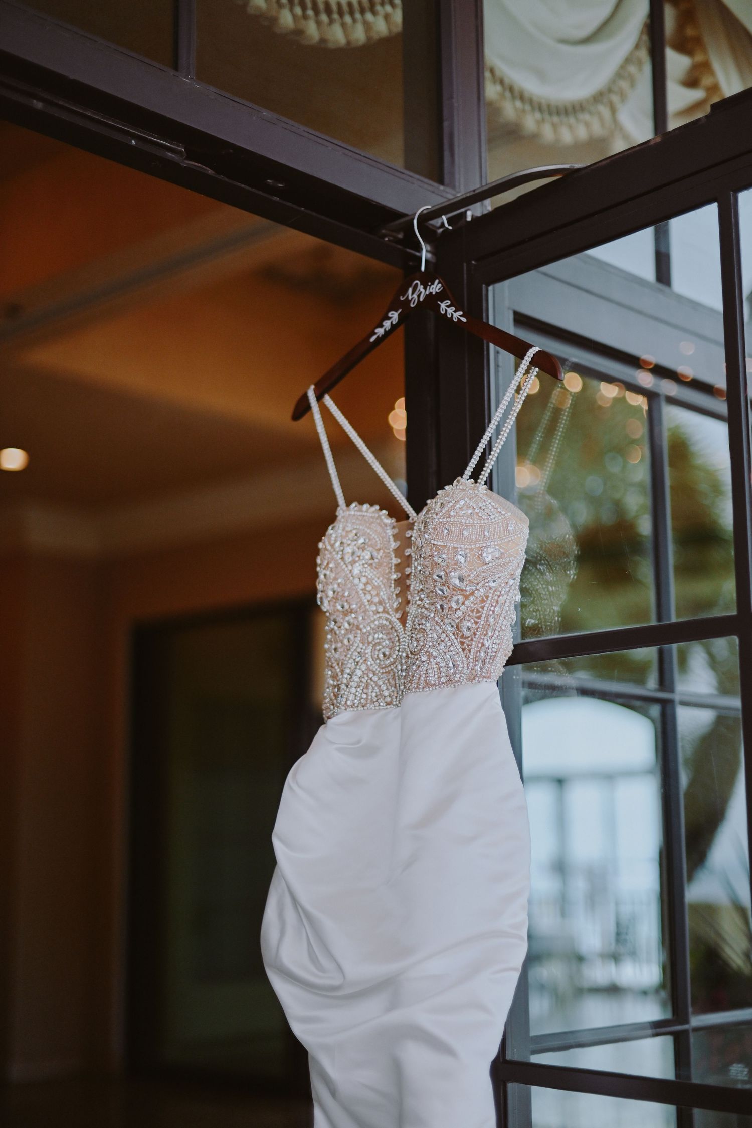 White beaded wedding dress with sweetheart neckline hanging on display near a window.