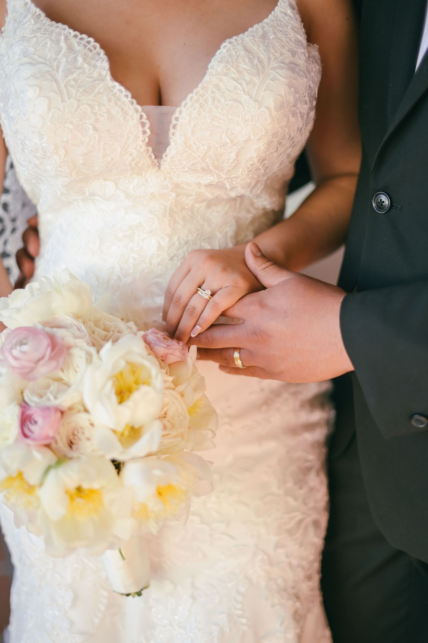 Close-up of wedding couple's hands exchanging rings with delicate lace dress and pink and white bouquet in view.