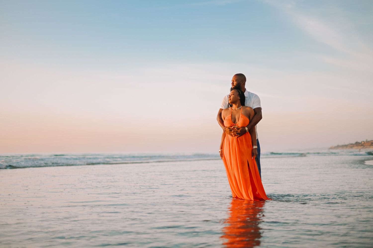 Woman in flowing orange dress stands at the edge of the ocean during sunset creating a stunning silhouette.