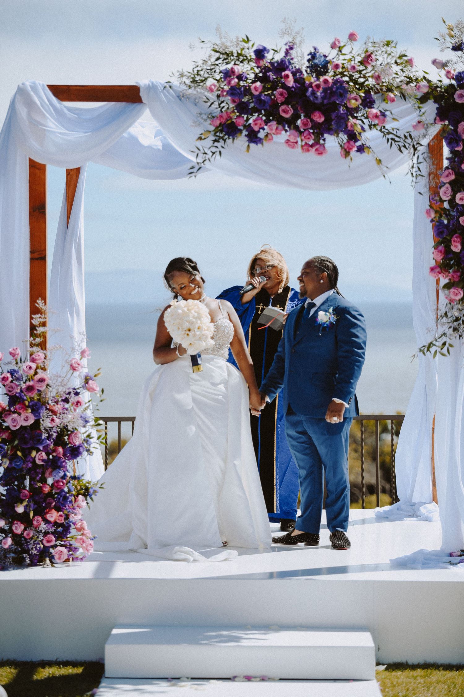 Wedding ceremony under white draped arch with purple and pink floral arrangements against ocean view.