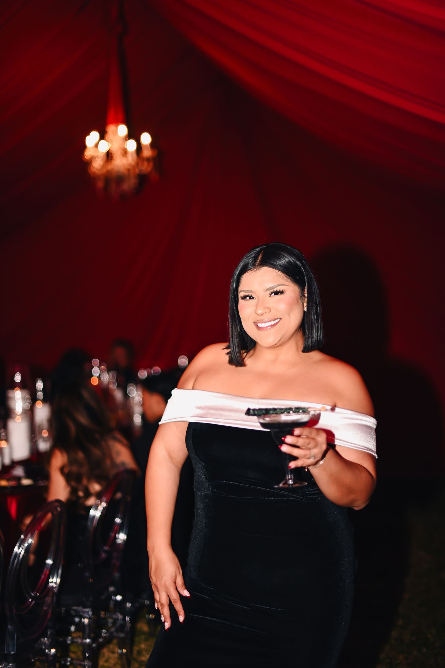 Woman in black dress holding cocktail at formal event with red decor.