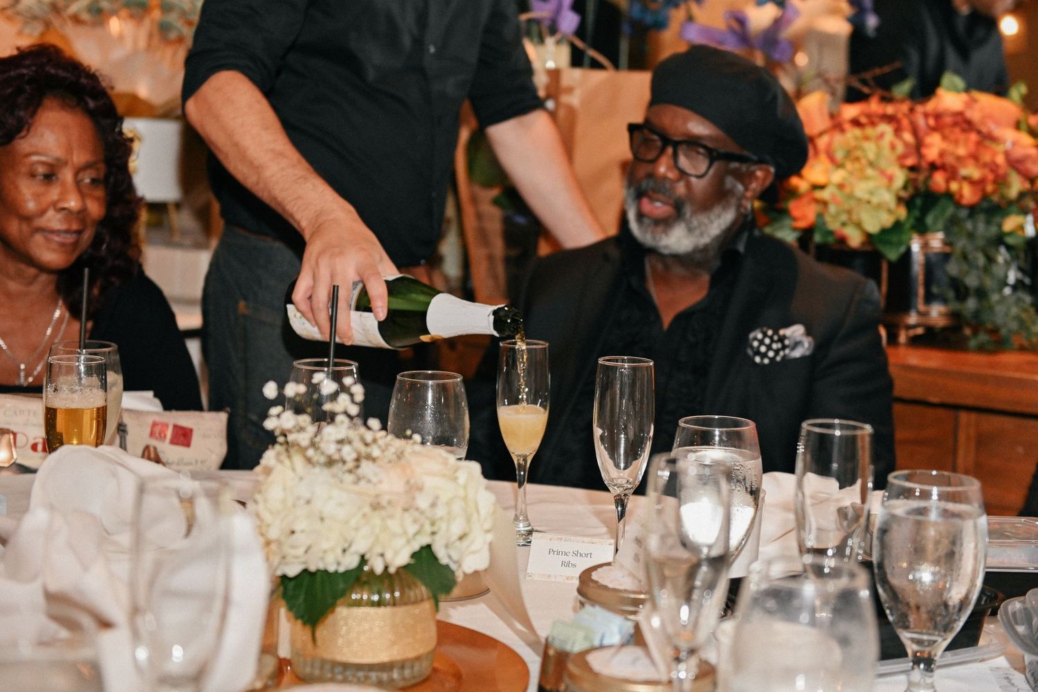 Guests seated at an elegant dinner table with floral centerpieces and champagne being served.