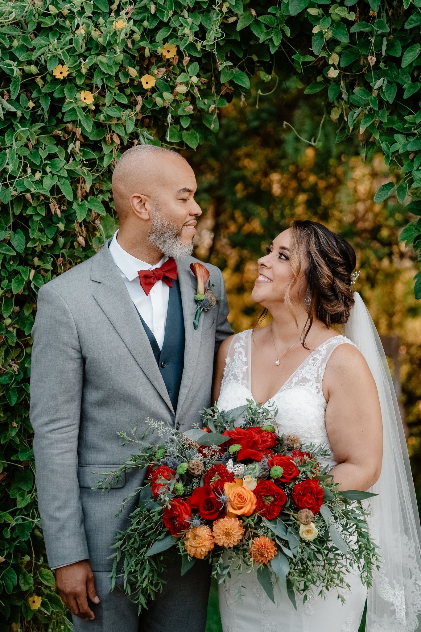 A bride and groom share a romantic moment against a backdrop of greenery with a vibrant red and orange bouquet.