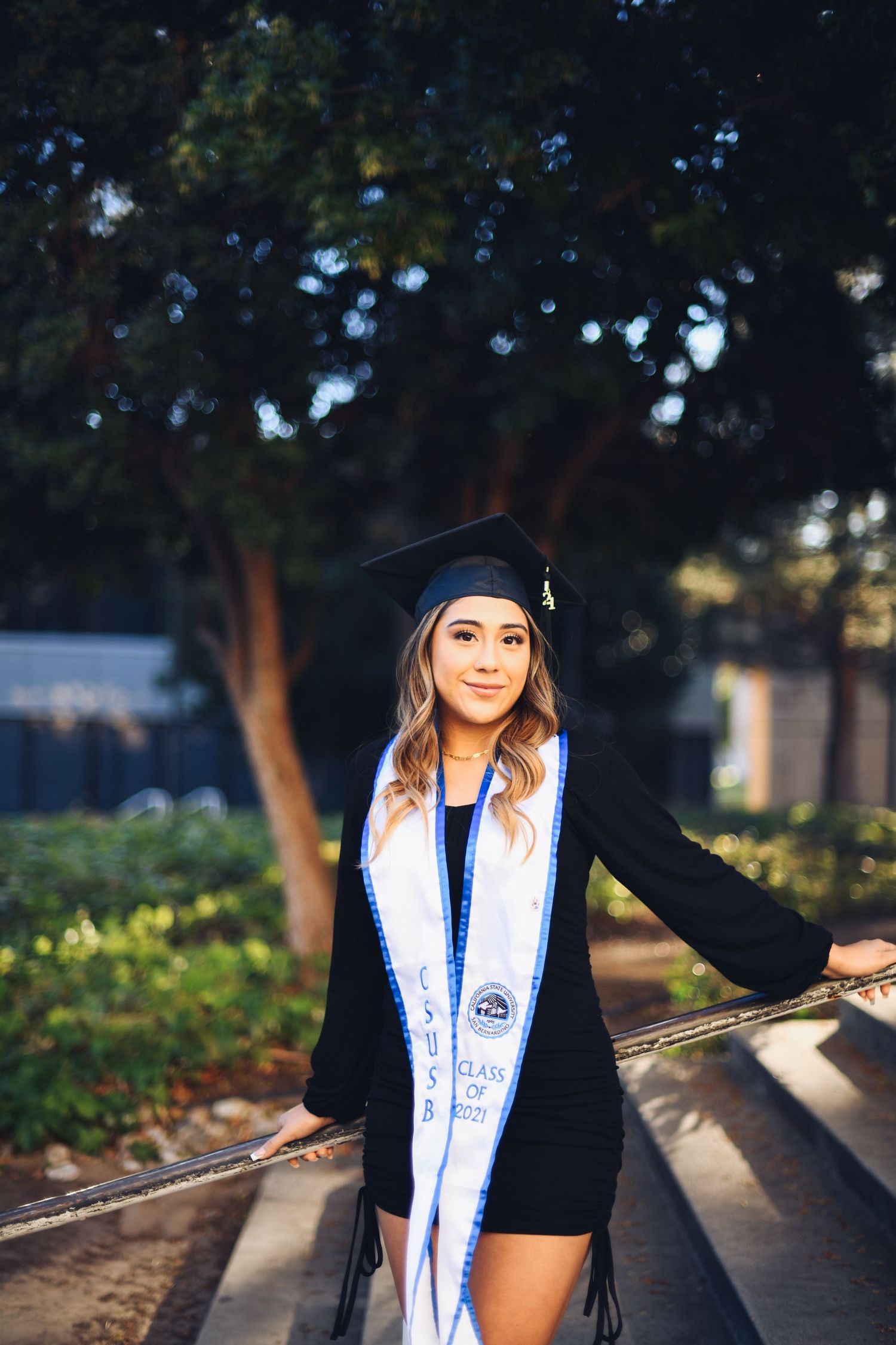 Graduate in black dress and white sash poses on steps outdoors under trees.
