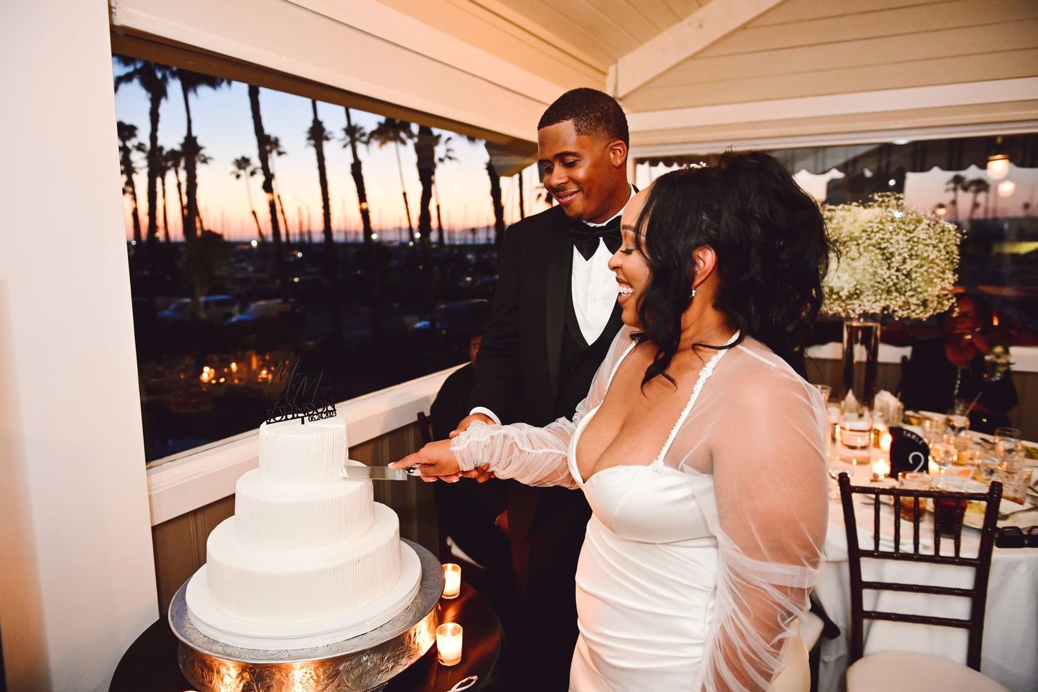 A couple cuts their elegant white wedding cake together at sunset with palm trees visible in the background.