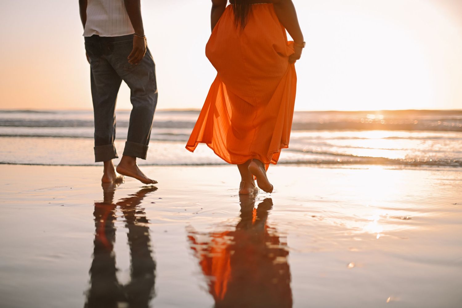 Silhouetted couple walking on beach at sunset with orange dress flowing in breeze.