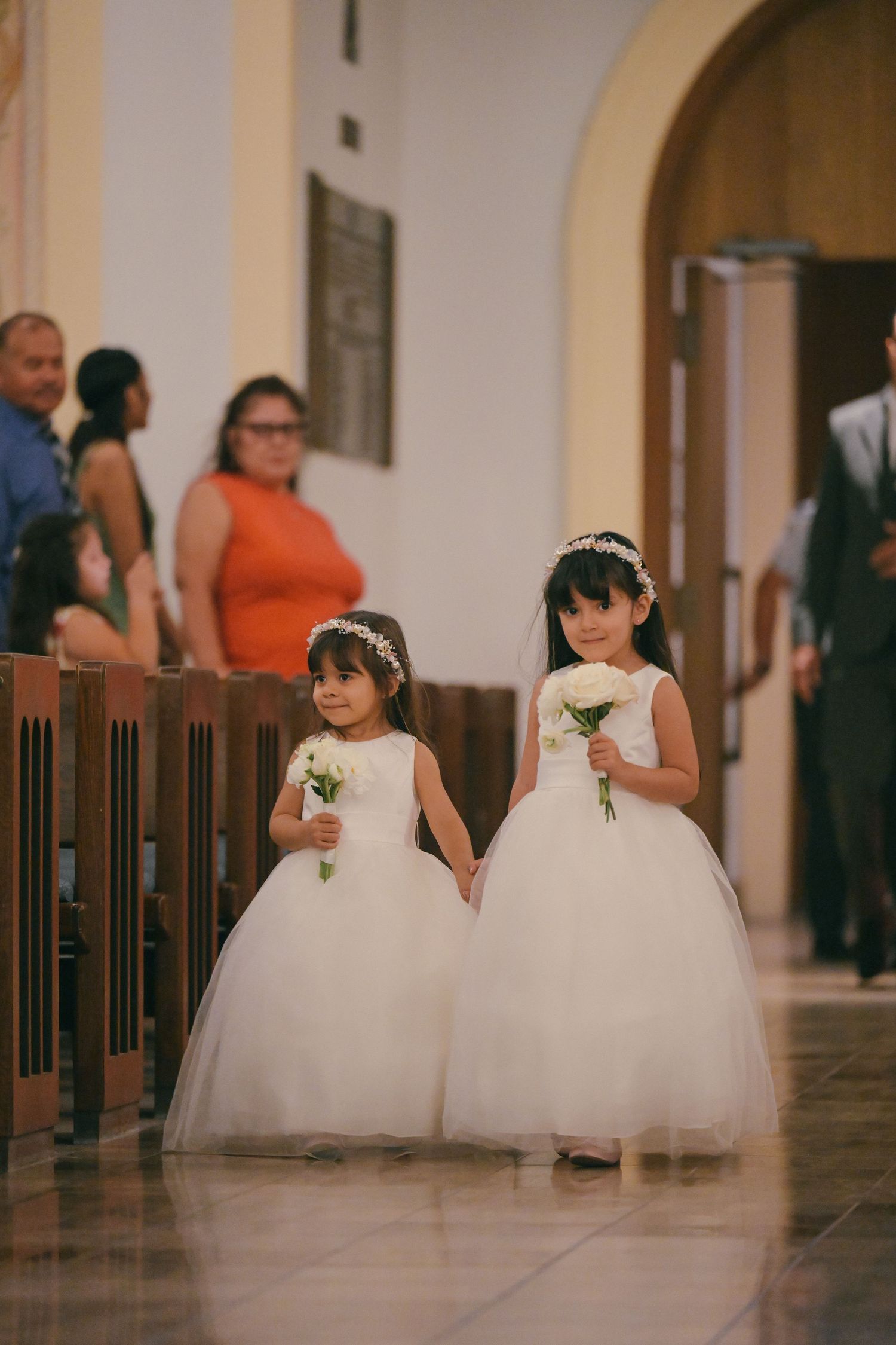 Two flower girls in white dresses walking down church aisle holding small bouquets.
