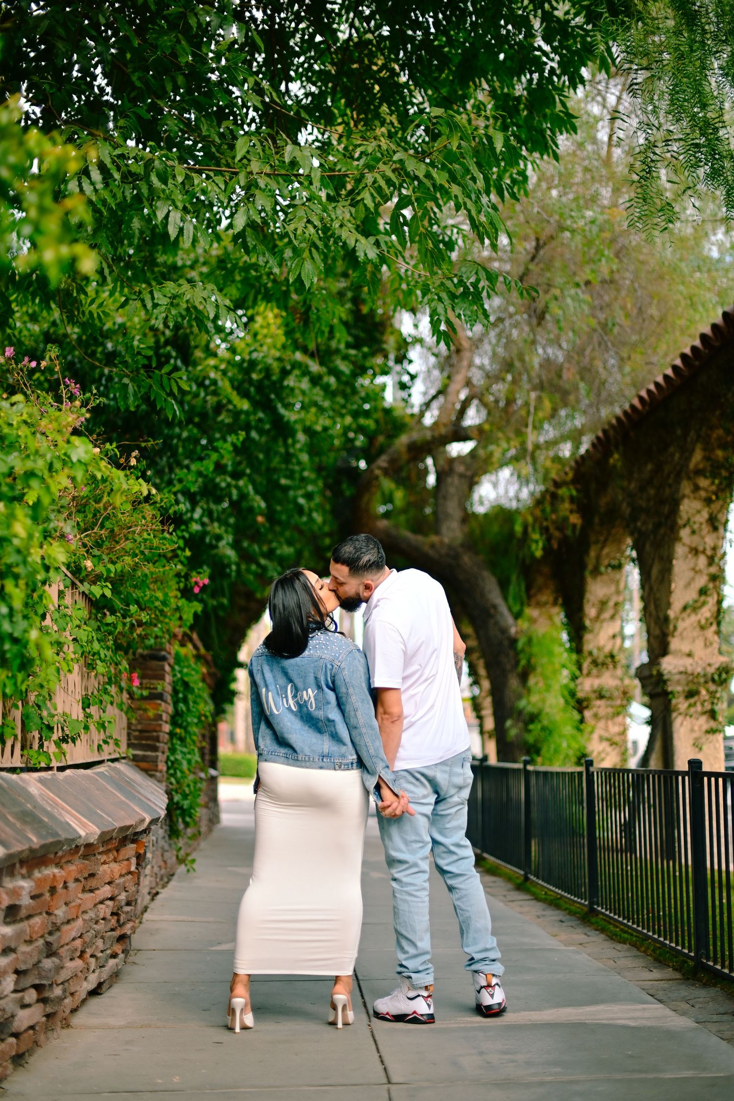 A romantic couple sharing a kiss on a tree-lined pathway with brick walls and iron railings.