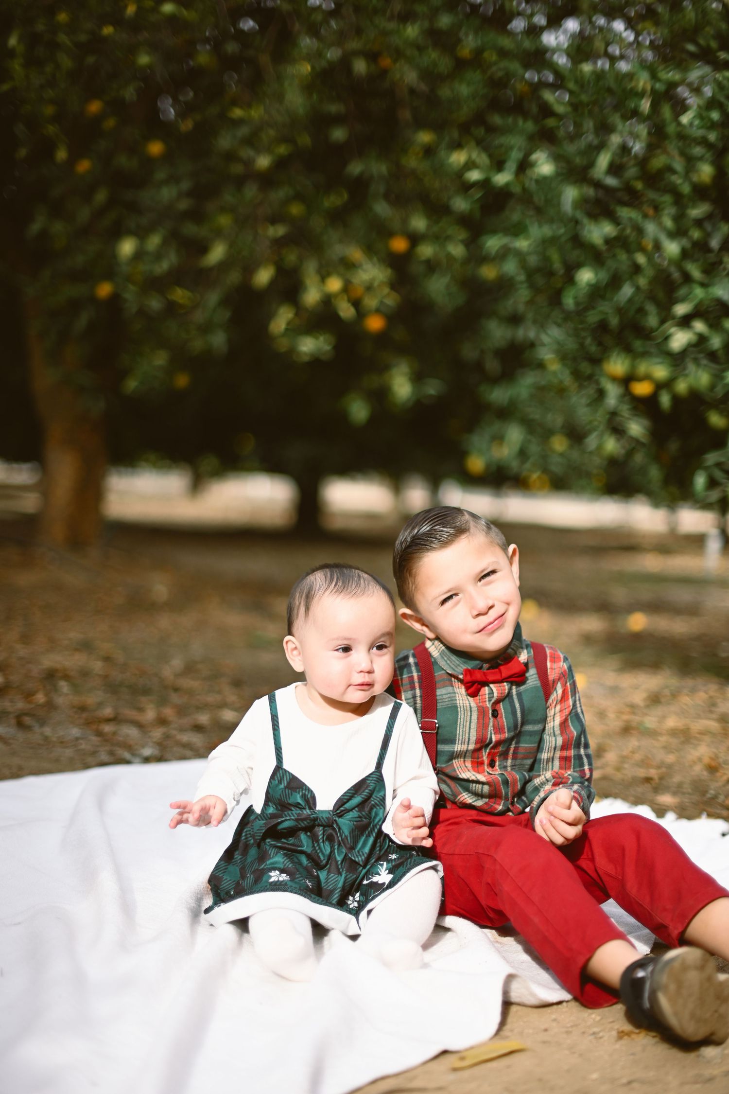 Two small children in vintage-style clothing sitting on a white blanket under autumn trees.