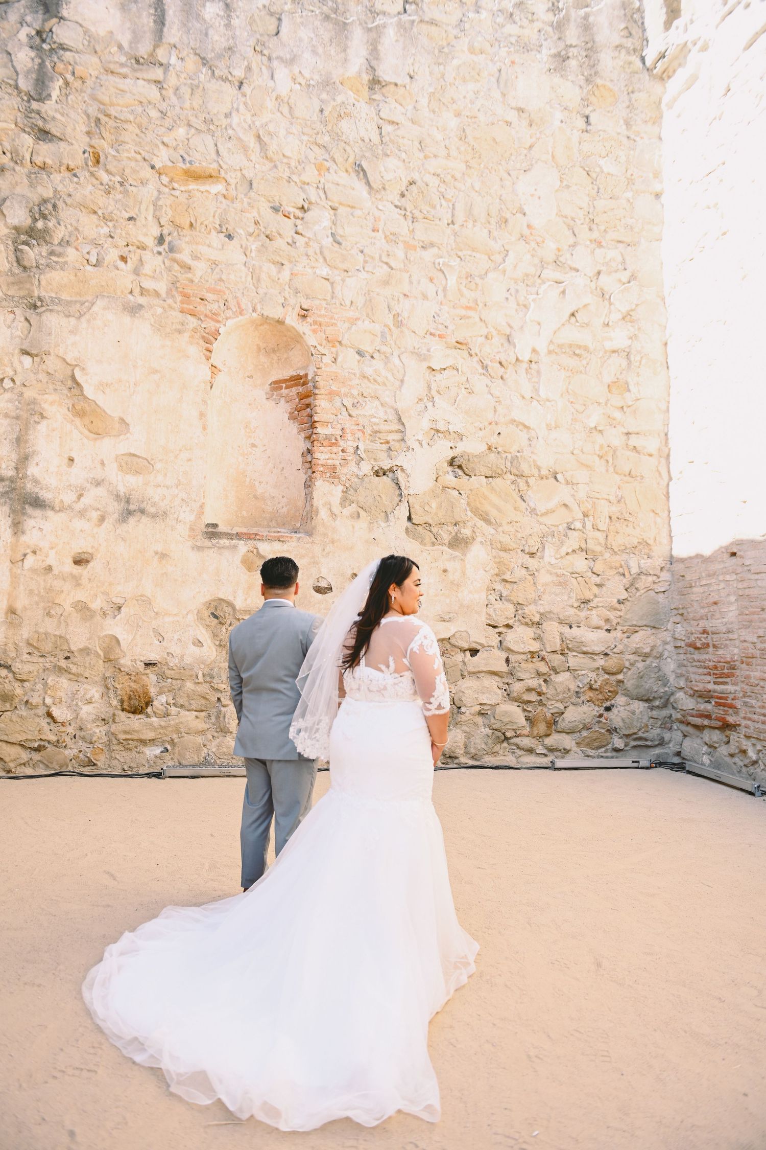 A romantic wedding couple standing against a rustic stone wall with flowing white dress in soft sunlight.