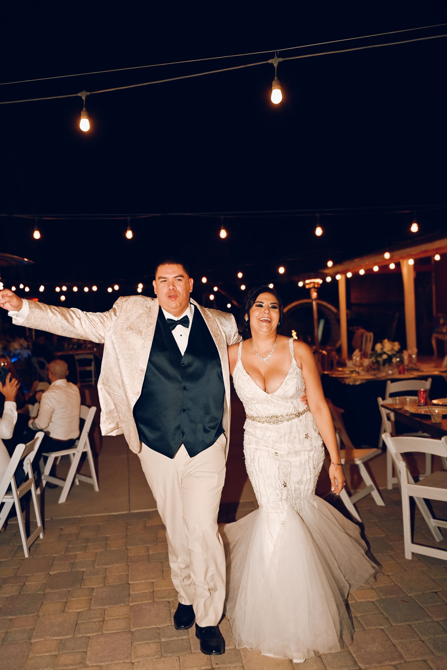Evening wedding reception outdoors with string lights and couple walking on patio.